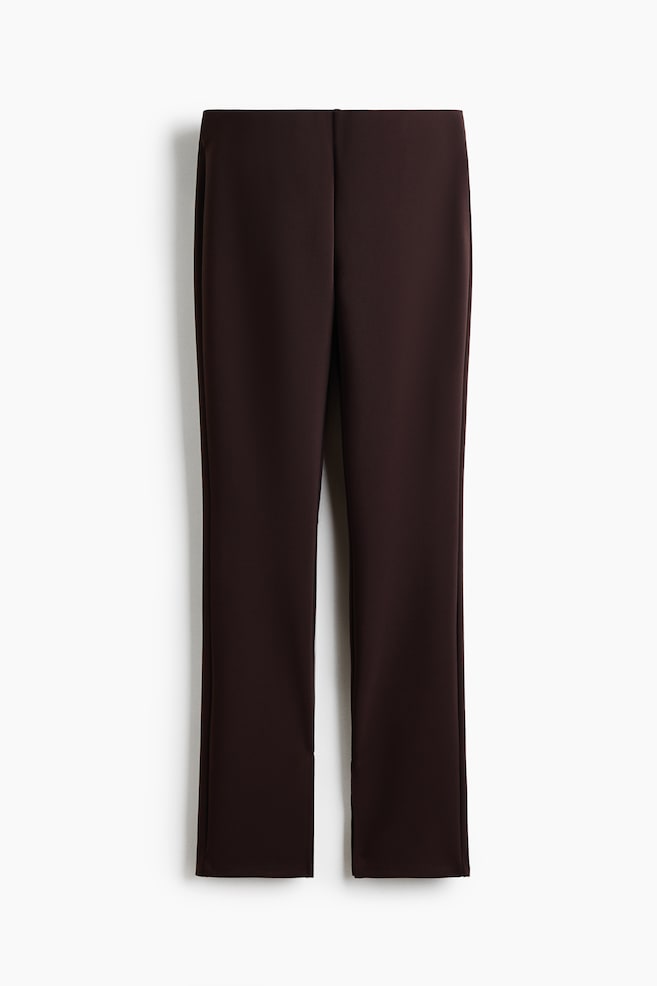 Dark brown trousers are positioned front-facing and laid flat, featuring a high-waisted design and a flat front. The trousers exhibit a straight leg cut with a visible centre crease running down each leg. - 1