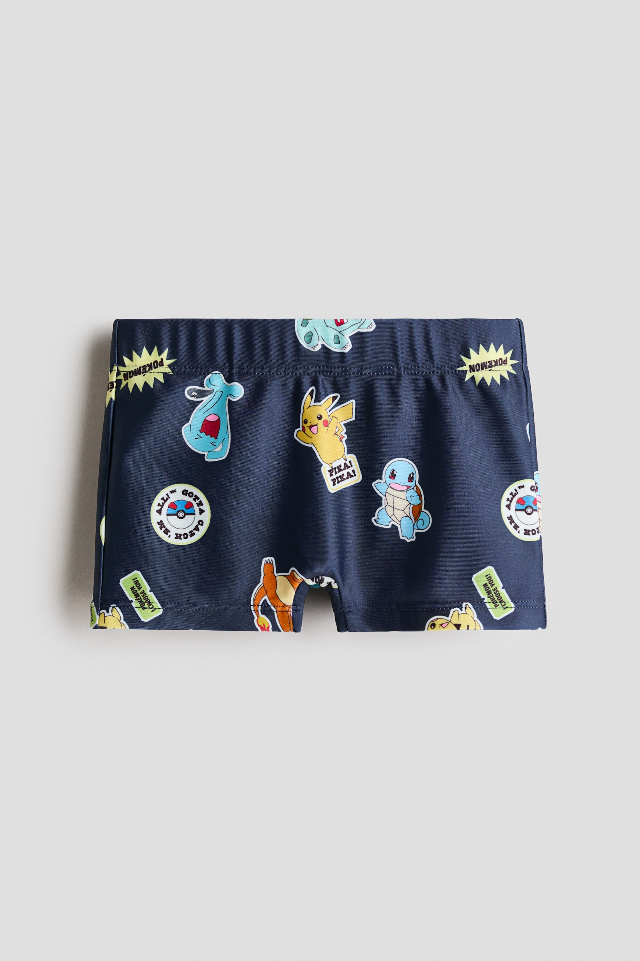 Grotere afbeelding bekijken: Een paar donkerblauwe zwembroeken ligt plat, met de voorkant zichtbaar, en heeft een all-over print van diverse kleurrijke Pokémon-personages en bijbehorende teksten. Een elastische tailleband vormt de bovenrand, en zichtbare stiksels omlijnen de korte beenopeningen en zijnaden.