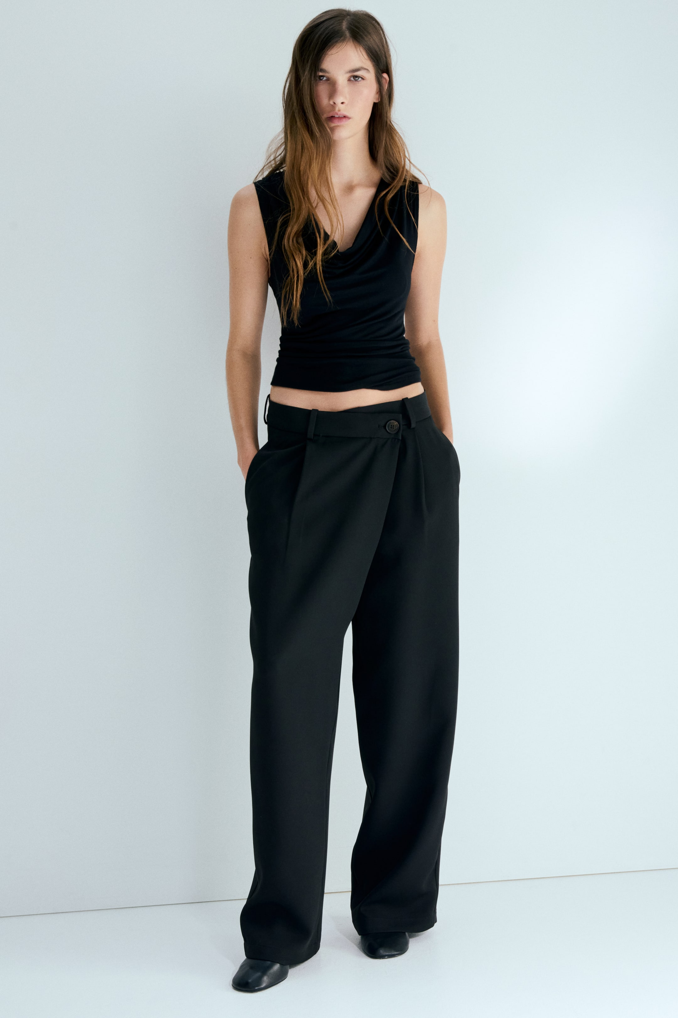 View larger image: Loose-fit, tailored trousers are being worn by the model, featuring a regular waist with an asymmetric wrapover front fastened by a visible button. The trousers showcase wide legs with pleats on the front, and the model is standing with hands in the pockets.