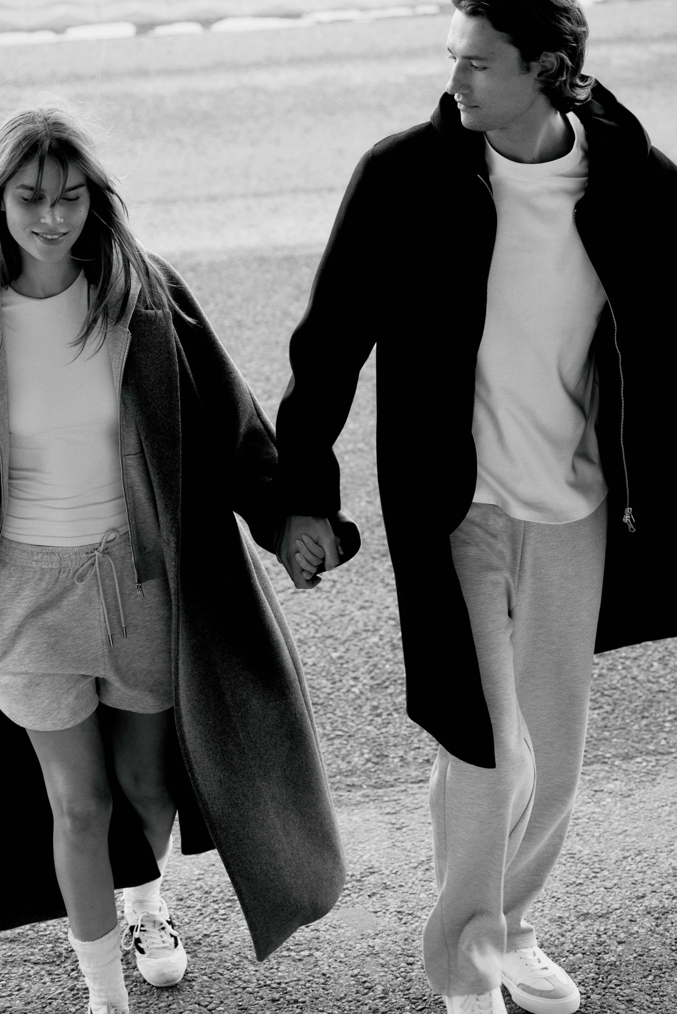 Büyük resmi görüntüle: Uzun siyah bir palto, hafif sol-ön açılı olarak yerleştirilmiş, rahat bir kesime ve kısmi ön fermuar kapamasına sahiptir. Altında beyaz bir bisiklet yaka tişört ve açık gri eşofman altı görünüyor.