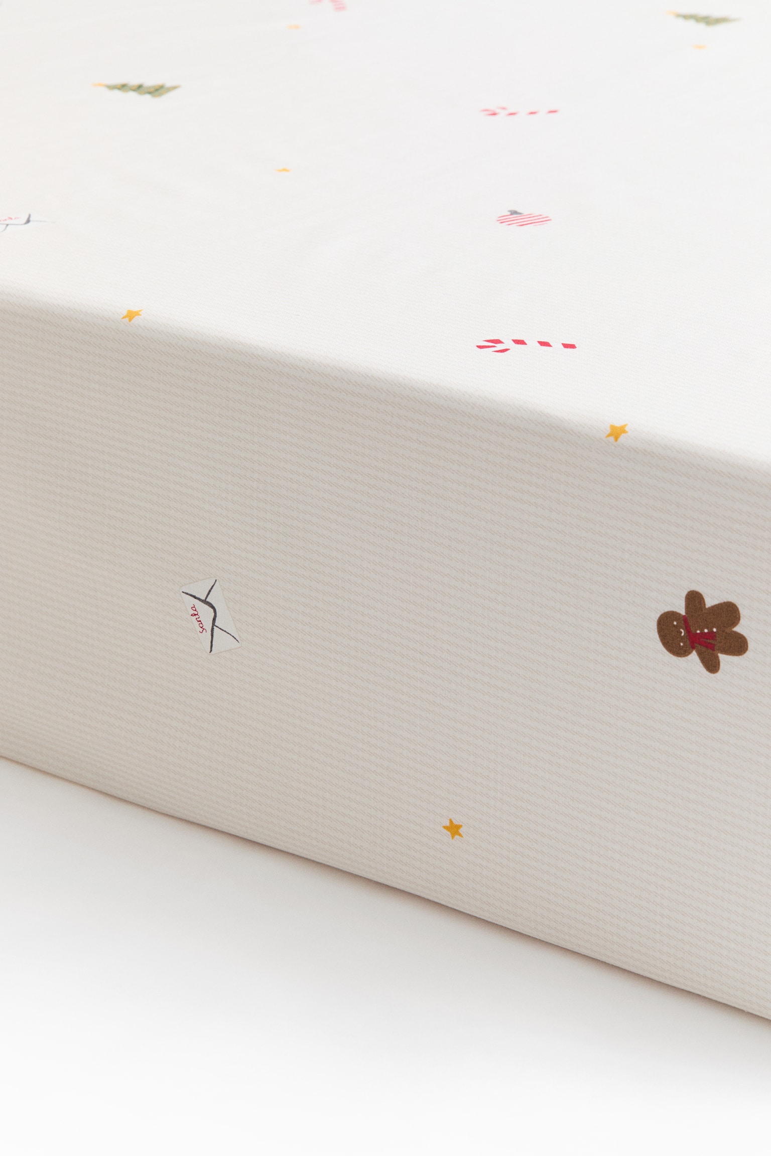 A white cotton fitted sheet covers the corner of a mattress, showing its snug fit over the top and side edges. The fabric features a scattered pattern of small illustrations, including gingerbread men, candy canes, envelopes, stars, and green trees.