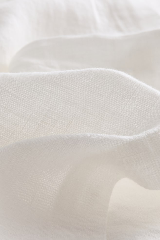 The white linen tablecloth fabric is presenting its characteristic weave and texture in a close-up view. It is gently draping, creating soft folds and subtle shadows. - 1
