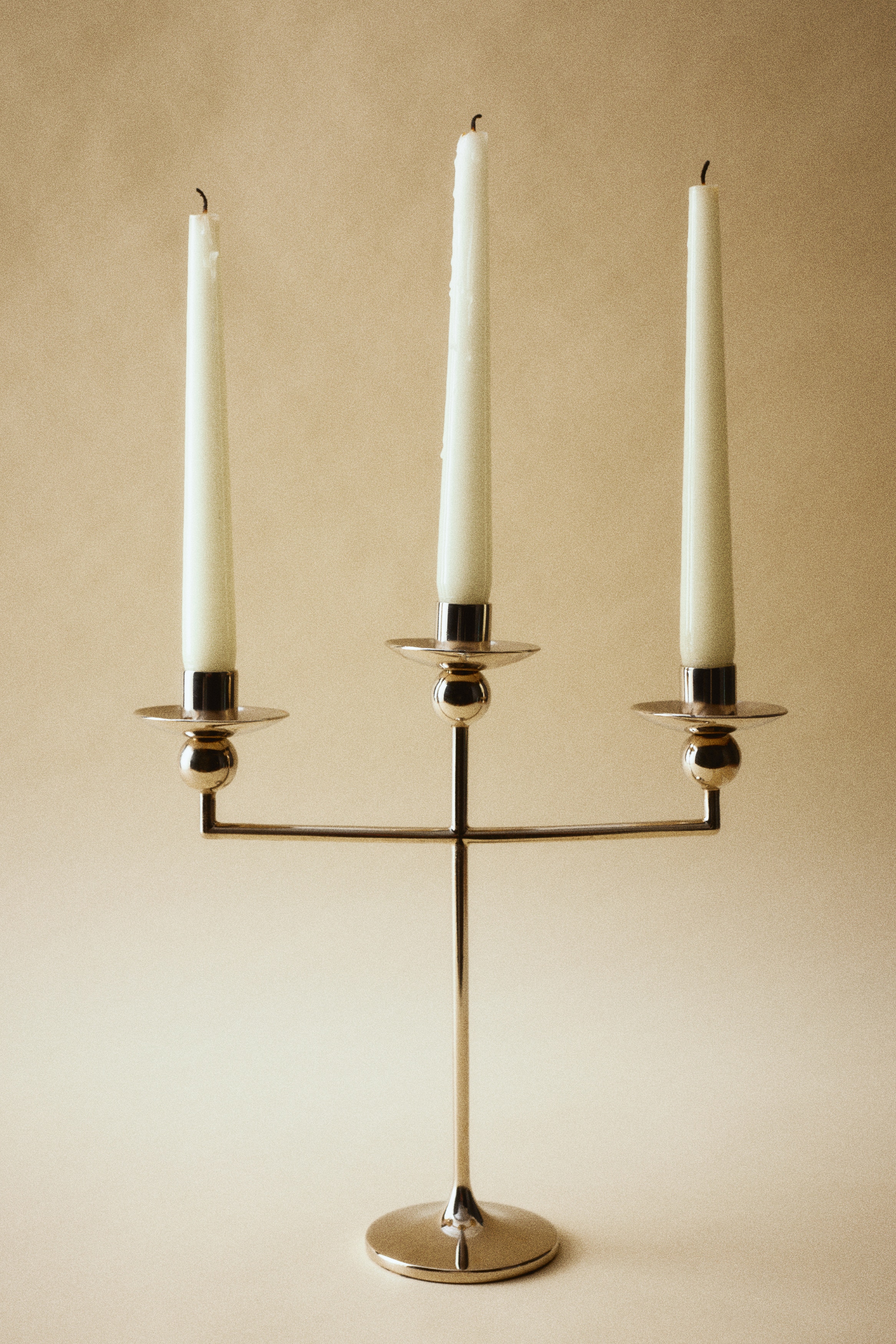 Un candelabru metalic cu trei braÈe stÄ drept, evidenÈiindu-Èi silueta elegantÄ Èi finisajul argintiu polisat din faÈÄ. Designul prezintÄ o tijÄ centralÄ subÈire care se ridicÄ dintr-o bazÄ circularÄ evazatÄ, susÈinÃ¢nd trei suporturi de lumÃ¢nÄri, fiecare cu un detaliu sferic sub o tÄviÈÄ puÈin adÃ¢ncÄ pentru cearÄ.