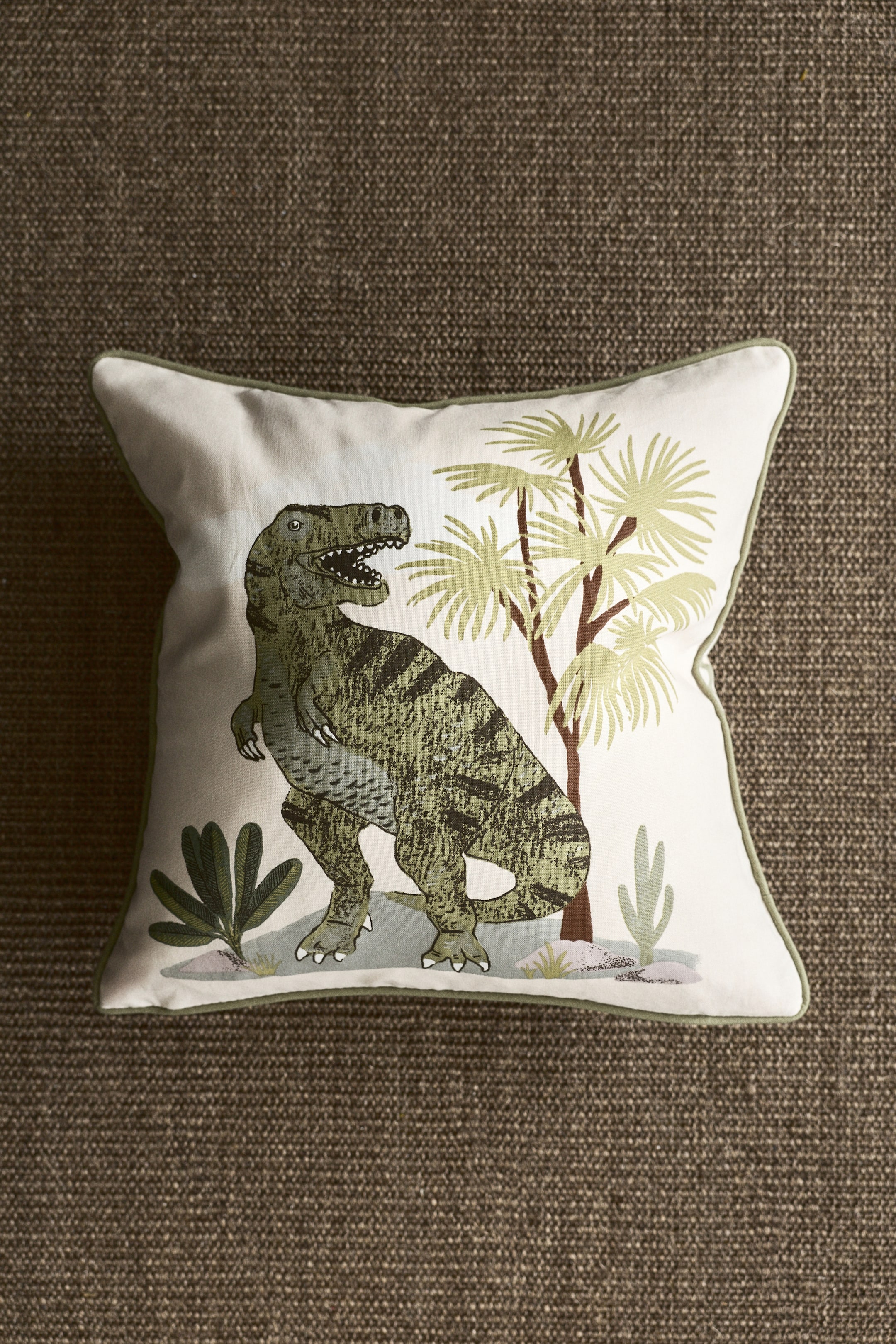 Visa större bild: En kudde ligger platt, något vinklad åt höger, med ett benvitt tyg och en central illustration av en grön Tyrannosaurus Rex. Illustrationen visar ljusgröna träd och mindre växter, och kudden är kantad med gröna passpoaler runt kanterna.