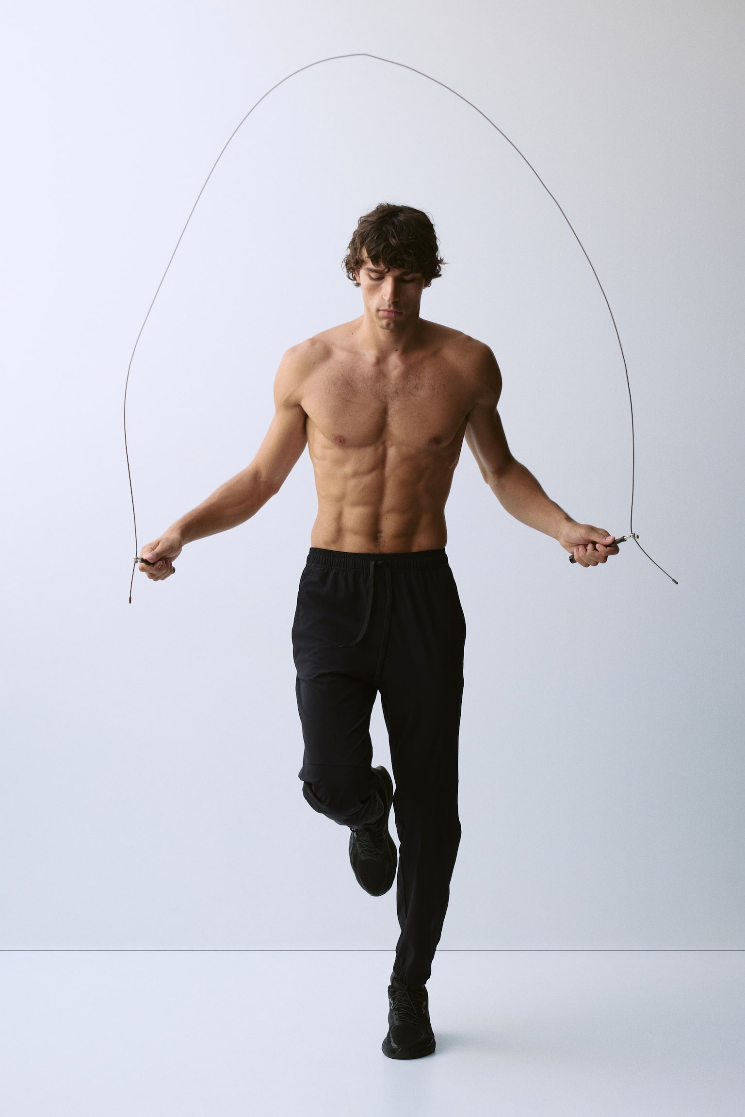 Black track trousers are shown from the front, featuring an elasticated waistband with a visible drawstring. Diagonal seams detail the upper leg, and elasticated cuffs display a side zip closure on one ankle. - 1