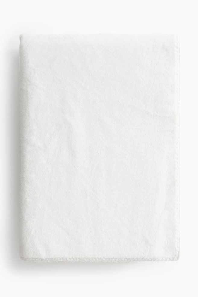 A soft pile fleece throw blanket in off-white is lying neatly folded in a rectangular shape, displaying its velvety texture and a whipstitched bottom hem. - 1