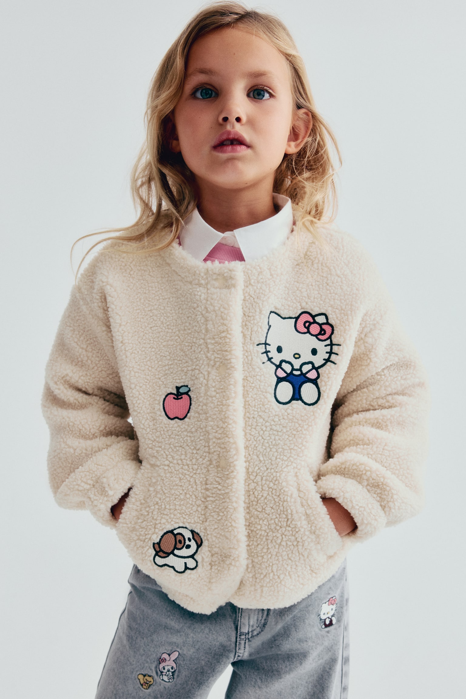 The Patch Detail Teddy Jacket is shown from the front, featuring a cream-coloured textured fleece material adorned with embroidered patches of Hello Kitty, a red apple, and a small dog. The collarless jacket is designed with a concealed front fastening and side slip pockets, and is layered over a white collared shirt. - 1