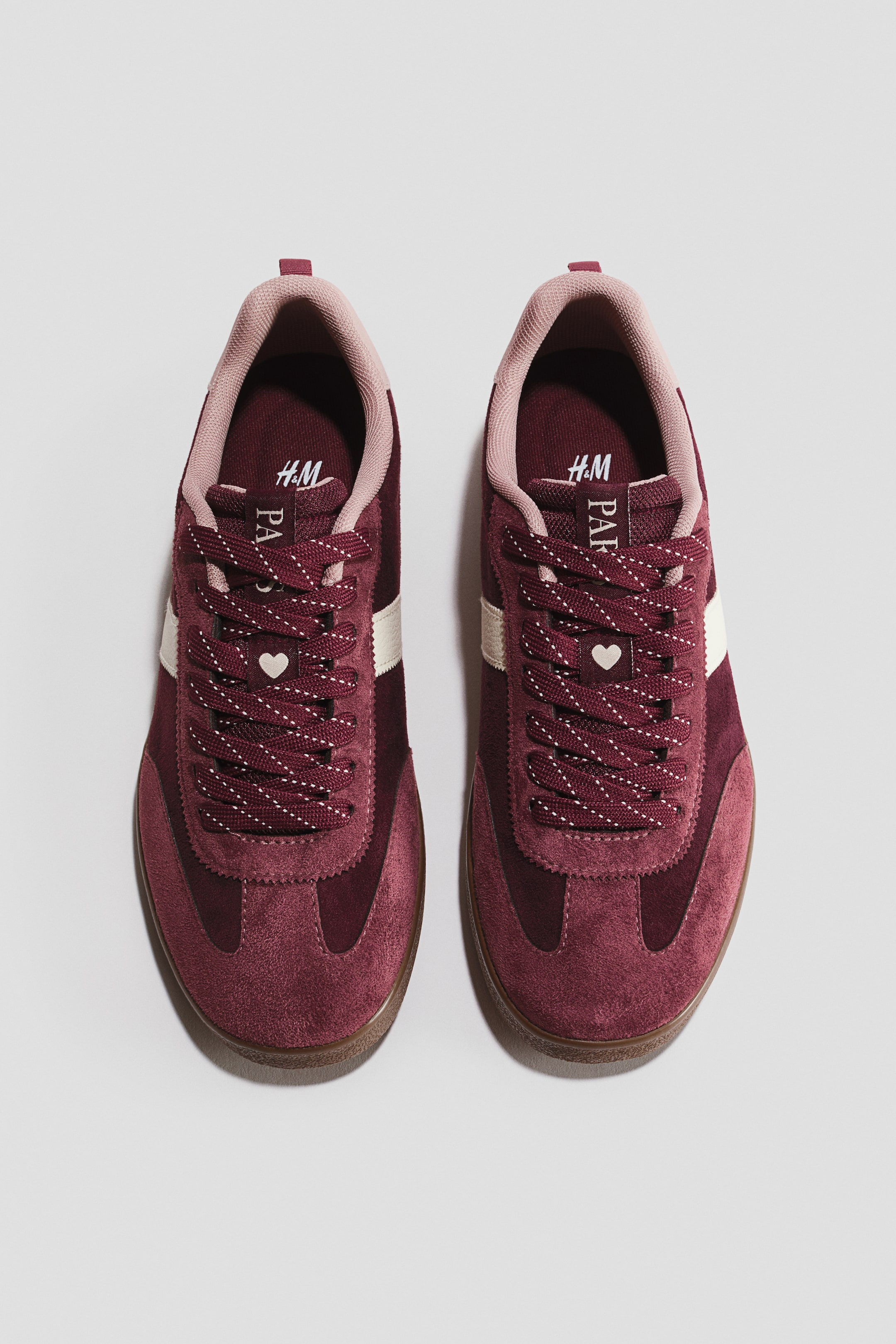 View larger image: A pair of burgundy suede sneakers features light pink lining, cream side stripes, and a brown gum sole. Speckled burgundy laces, adorned with a small white heart charm, tie the shoes, and "H&M" and "PAF" branding is visible on the tongue.