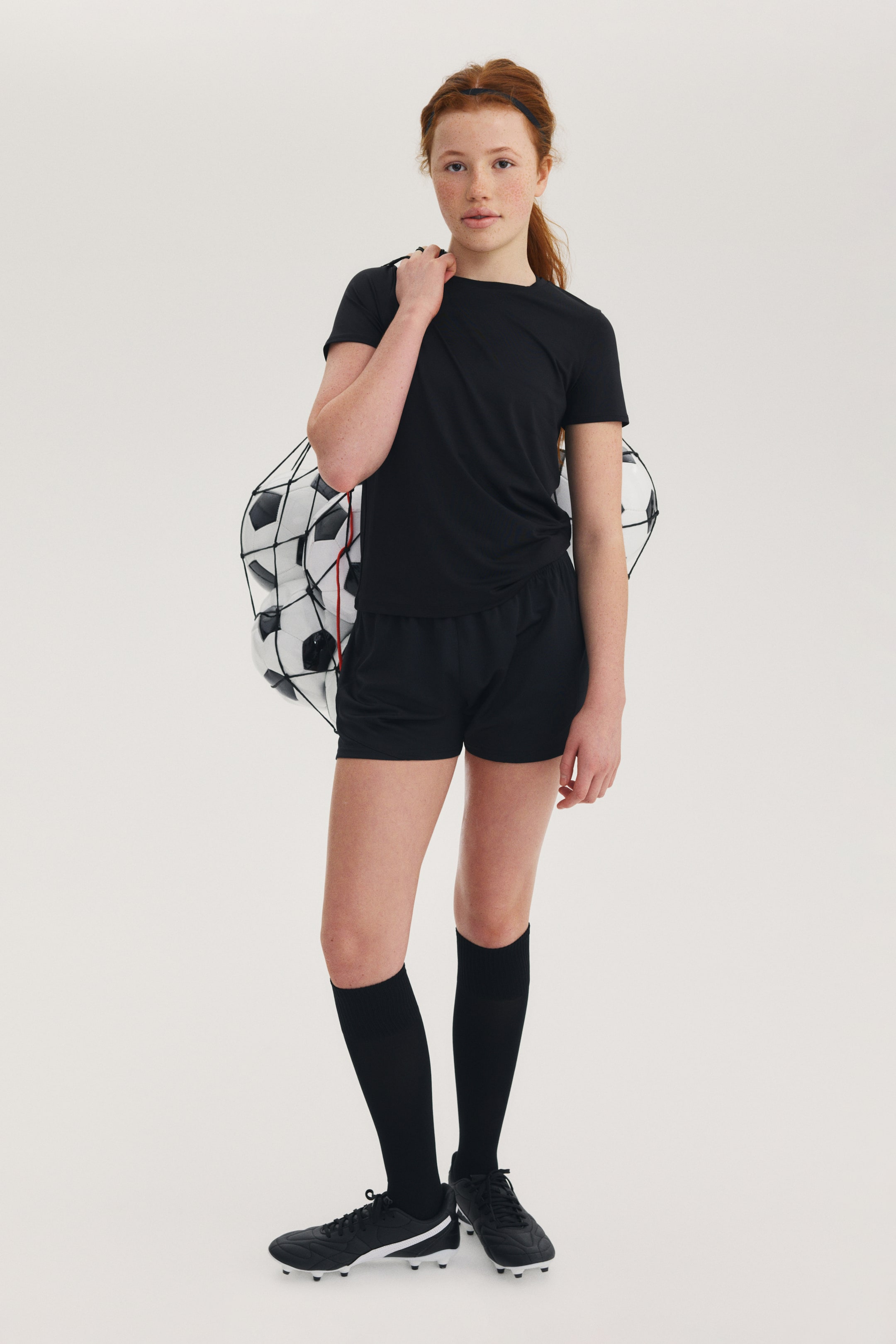 Grotere afbeelding bekijken: Er wordt een zwart sport-T-shirt met korte mouwen en een ronde hals gedragen, gecombineerd met zwarte sportshorts met een elastische tailleband. Zwarte kniehoge sokken en zwarte voetbalschoenen met witte accenten maken de outfit compleet, terwijl een zwarte nettas met zwarte en witte voetballen over de schouder wordt gedragen.