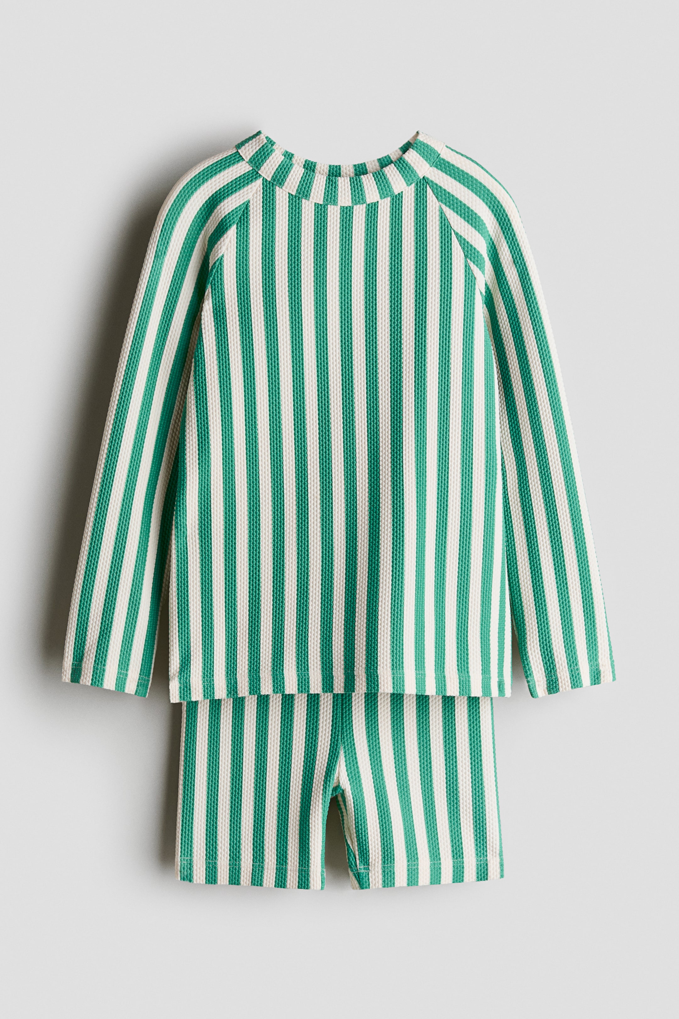 Visa större bild: H&M Adorables badset innehåller en badtopp och ett par badshorts, båda med ett grönt och benvitt vertikalt randmönster på texturerat tyg. Toppen är designad med en rund halsringning och långa raglanärmar, medan de matchande shortsen har korta ben.