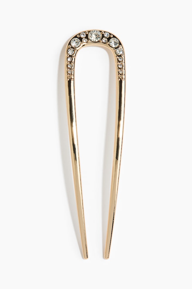 Een haarspeld in goudtint, U-vormig, is van voren weergegeven en toont een gebogen bovendeel versierd met heldere, ronde rhinestones in verschillende formaten. De speld loopt taps toe in twee lange, gladde pinnen met een hoogglans afwerking. - 1