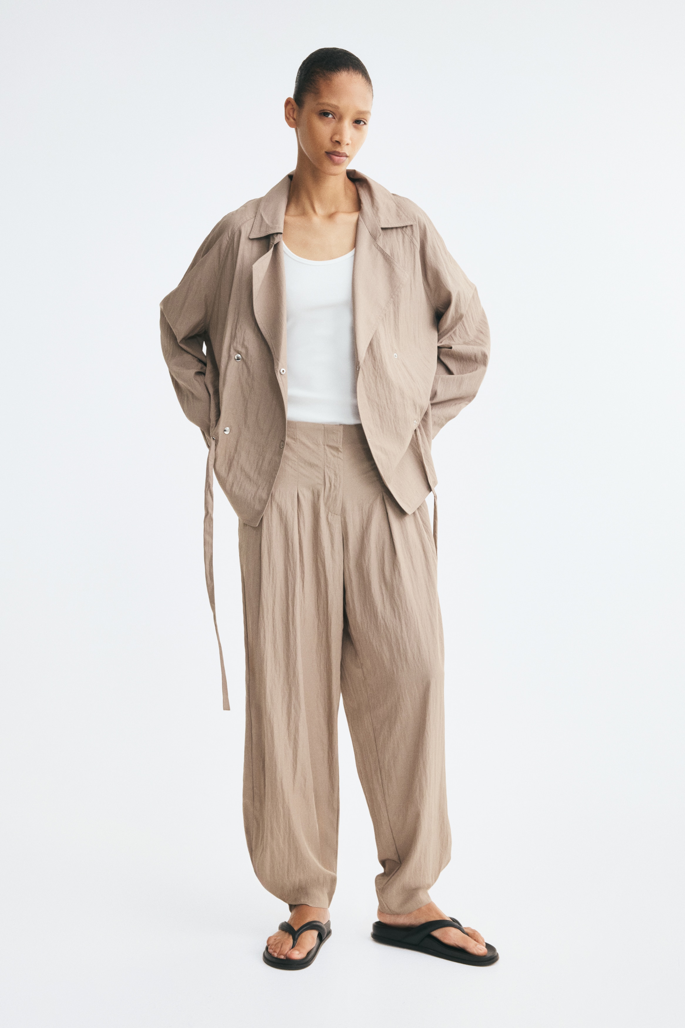 Taupe broek heeft een brede, hoog getailleerde band, waaruit diepe plooien langs de voorkant naar beneden vloeien. De broek vertoont een gekreukte textuur en een knoopsluiting voltooit de taille aan de voorkant.