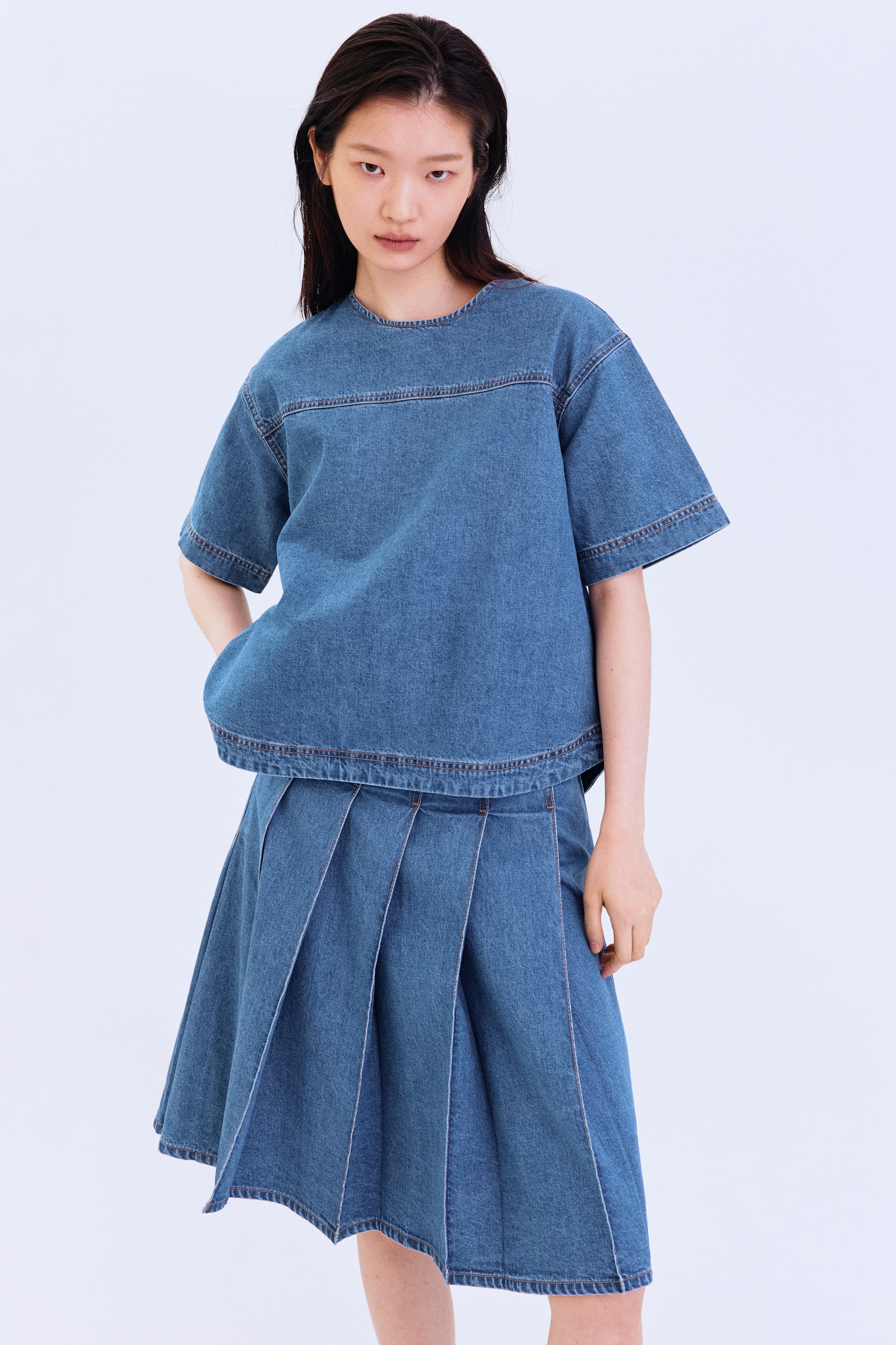 View larger image: Presenting a medium blue denim short-sleeved top and pleated skirt set, displayed front-facing. The top boasts a crew neck, a boxy fit, and a horizontal seam across the chest. The knee-length skirt features an A-line silhouette with visible pleats, all highlighted by contrasting stitching.