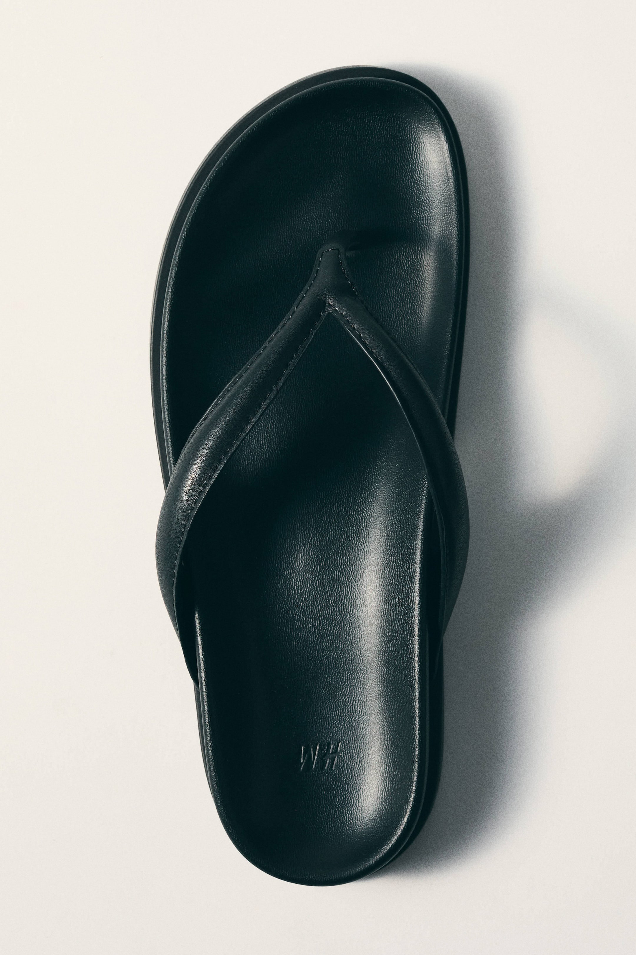 View larger image: A single black flip-flop sandal is lying flat, seen from a top-down view, featuring a smooth, slightly textured footbed with an embossed "H&M" logo near the heel and two rounded straps forming a V-shape.
