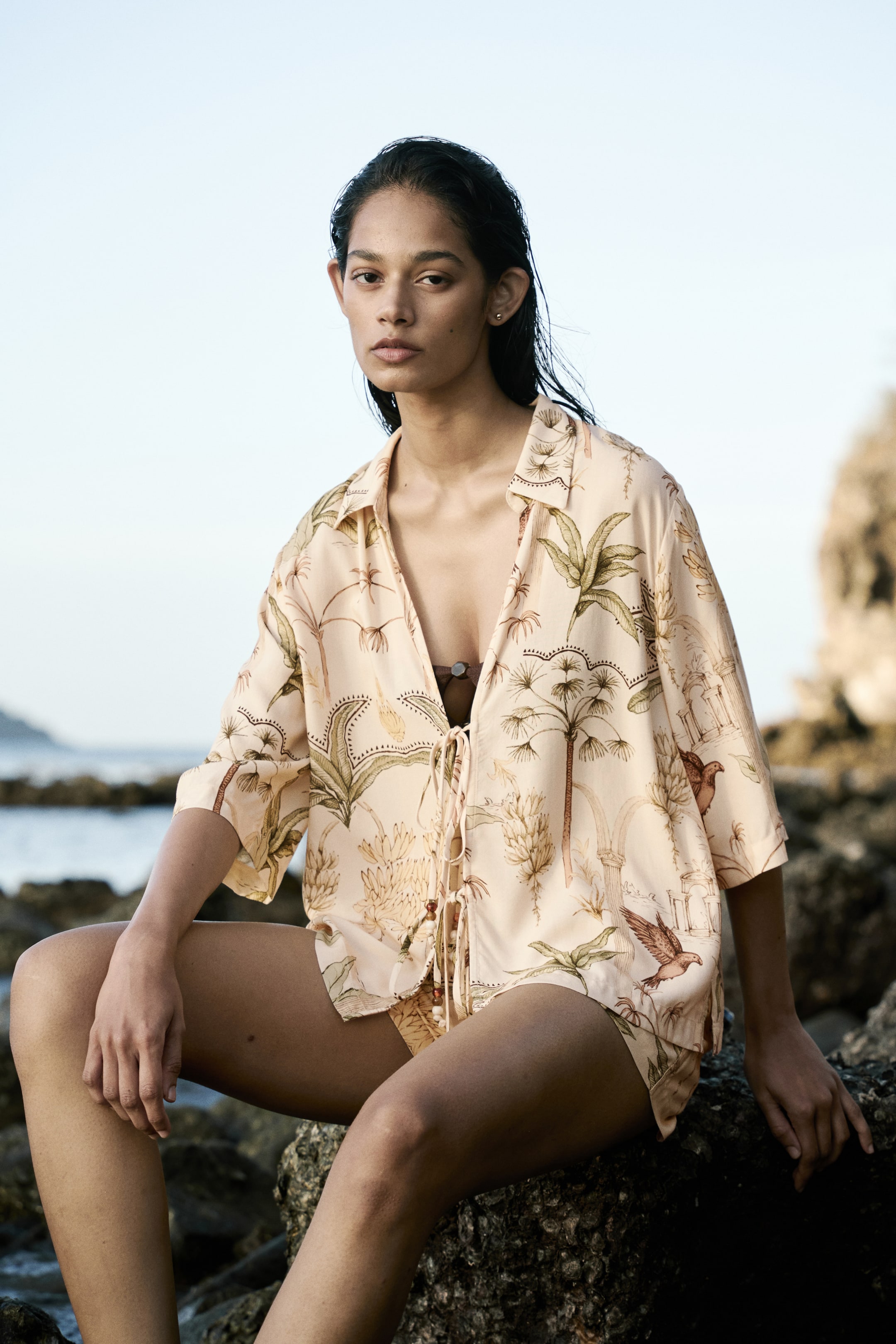 View larger image: The model dons a loose-fit shirt crafted from a viscose weave, showcasing a vibrant tropical print. It boasts a collar, wide elbow-length sleeves, and a straight-cut hem. The front is fastened with two sets of bead-embellished ties, subtly revealing a dark bikini top underneath.