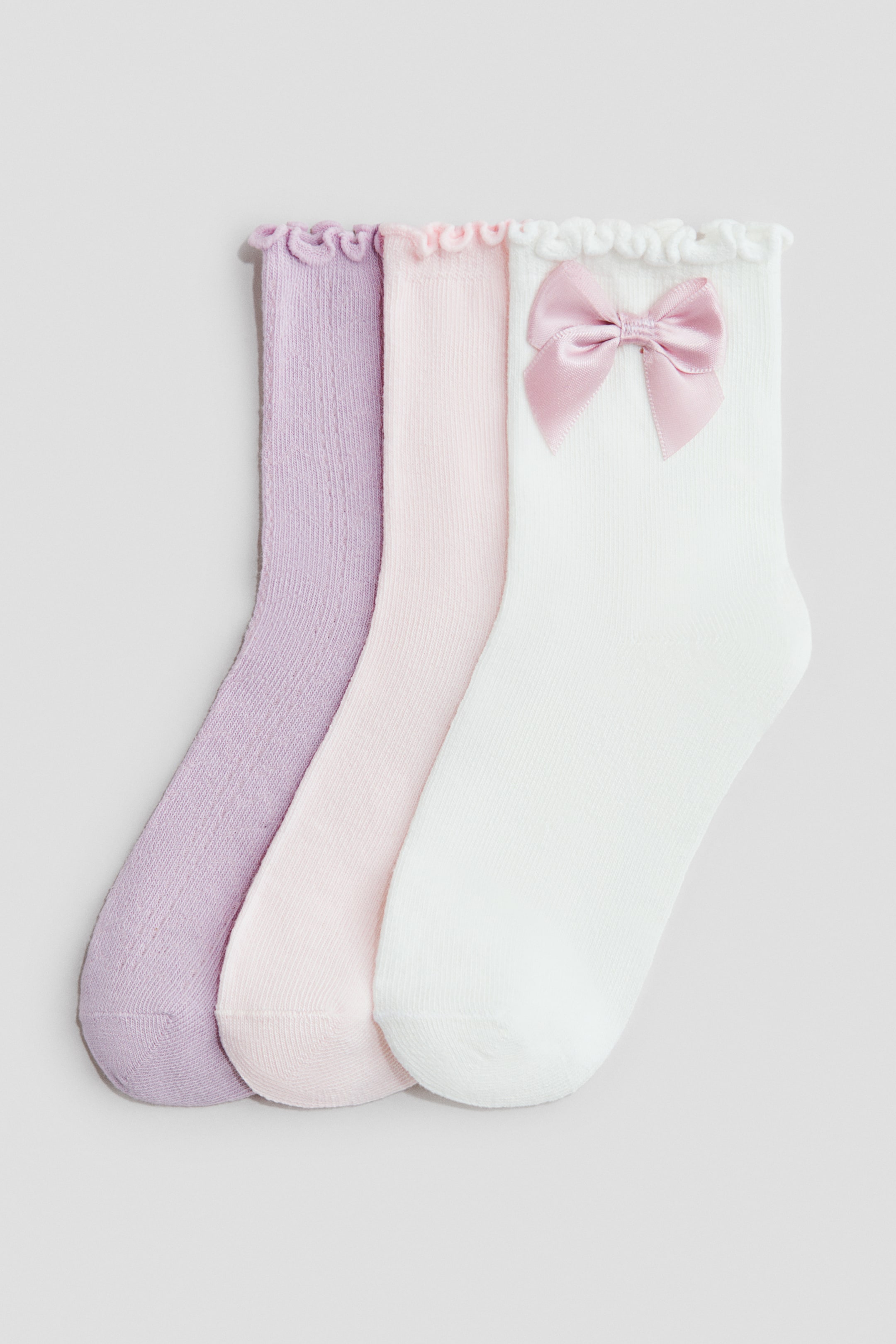 View larger image: Three fine-knit socks are shown from the front, showcasing elasticated, frill-trimmed tops. They come in lavender, light pink, and white, with the white sock adorned with a pink satin bow.