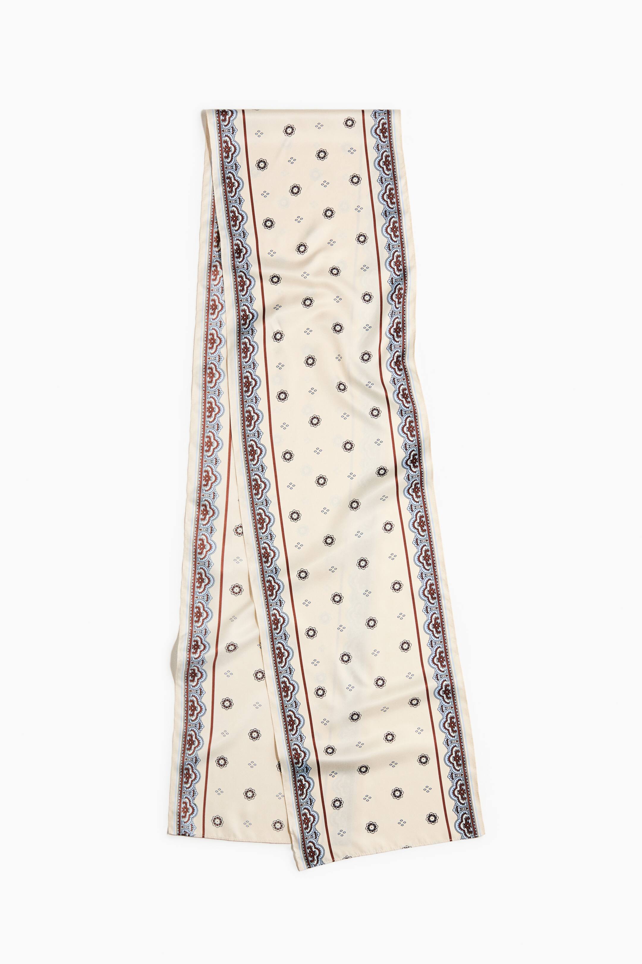 הצגת תמונה גדולה יותר: צעיף סאטן ארוך מציג רקע בצבע קרם המעוטר בדוגמה חוזרת של מוטיבים עגולים קטנים וצורות יהלום זעירות. הוא מתאפיין בשני שוליים אנכיים מובחנים, שכל אחד מהם מורכב מקווים בגוון חום-אדמומי המסגירים עיטור בצורת גלילה בצבעי כחול וחום-אדמומי.