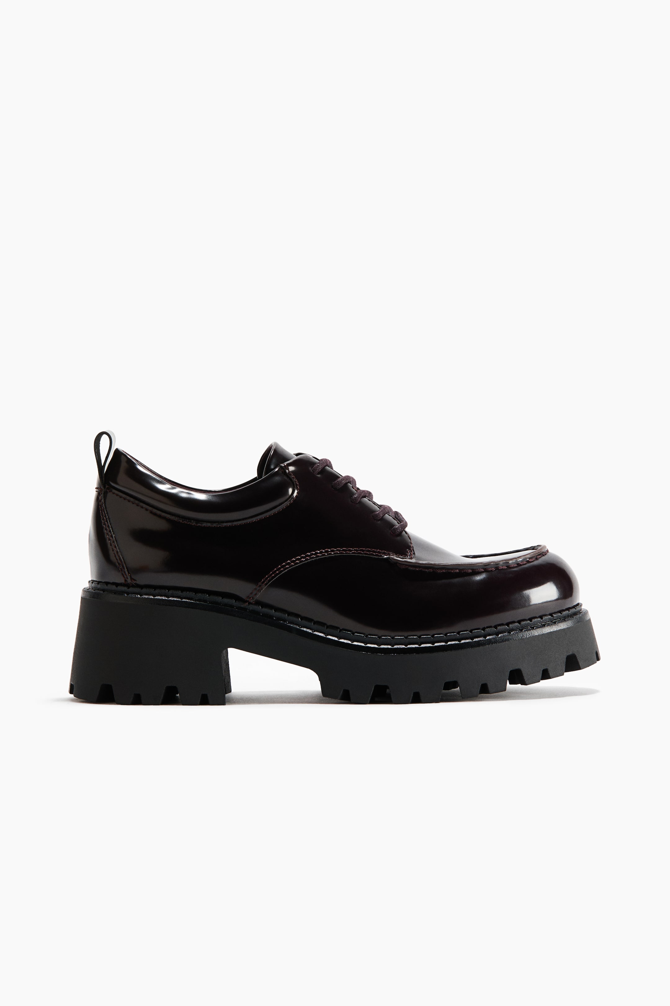 View larger image: A dark burgundy patent leather lace-up shoe is shown in side profile, featuring a moc-toe design, dark laces, and a small pull tab at the heel collar. It rests on a chunky black lug sole with a prominent block heel and white contrast stitching around the welt.