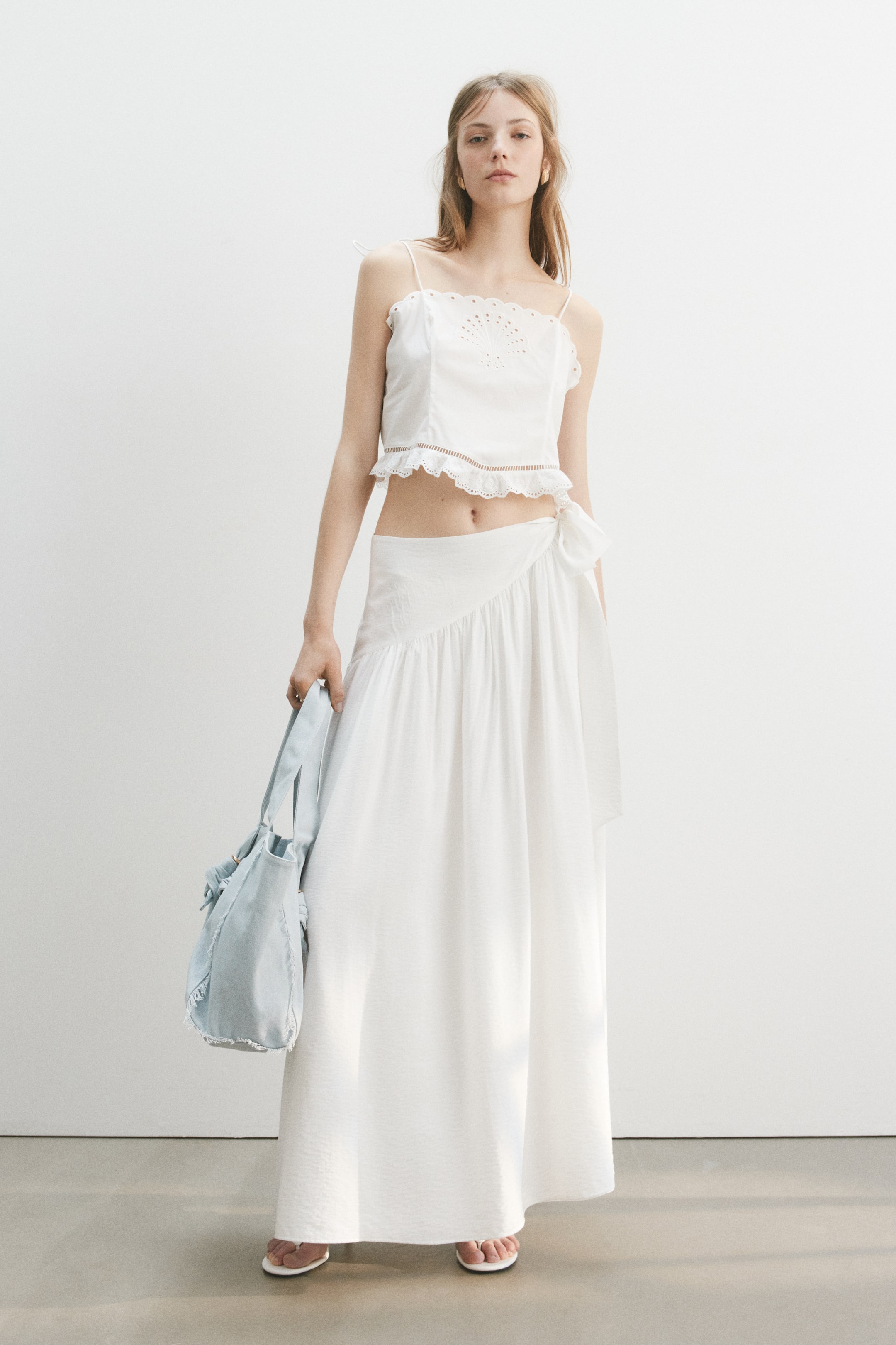 View larger image: A white cropped camisole top featuring thin spaghetti straps, a scalloped neckline with eyelet embroidery, and a ruffled hem, while a white maxi wrap skirt ties at the left hip with a bow and flows down with gathers, both presented from a front-facing angle.