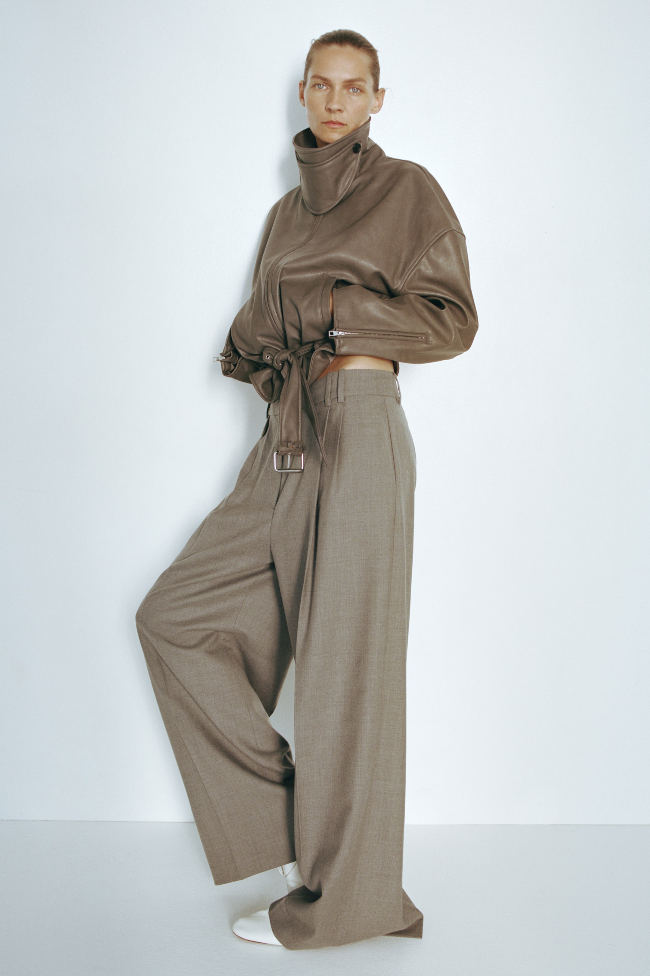 View larger image: Loose-fit, wide-leg trousers in a taupe woven fabric are being worn by the model, featuring a high waist, front pleats and pressed creases. The trousers are styled with a cropped, high-collar brown leather jacket and white shoes.