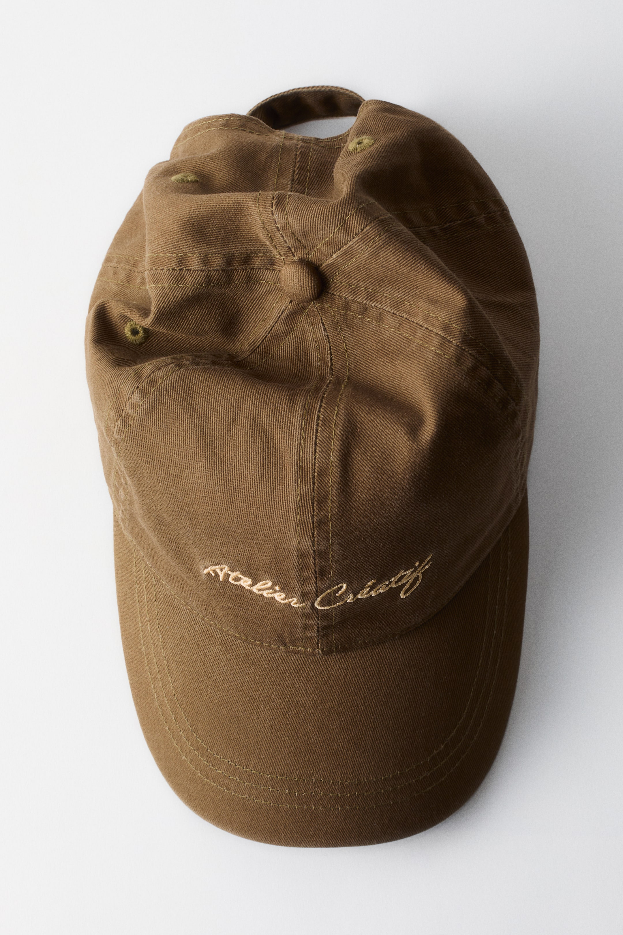 View larger image: A brown baseball cap lies flat, showcasing a six-panel crown topped with a fabric-covered button and featuring embroidered eyelets. The curved brim displays multiple rows of stitching, and light-colored text reading "Atelier Créatif" is embroidered on the front panel.
