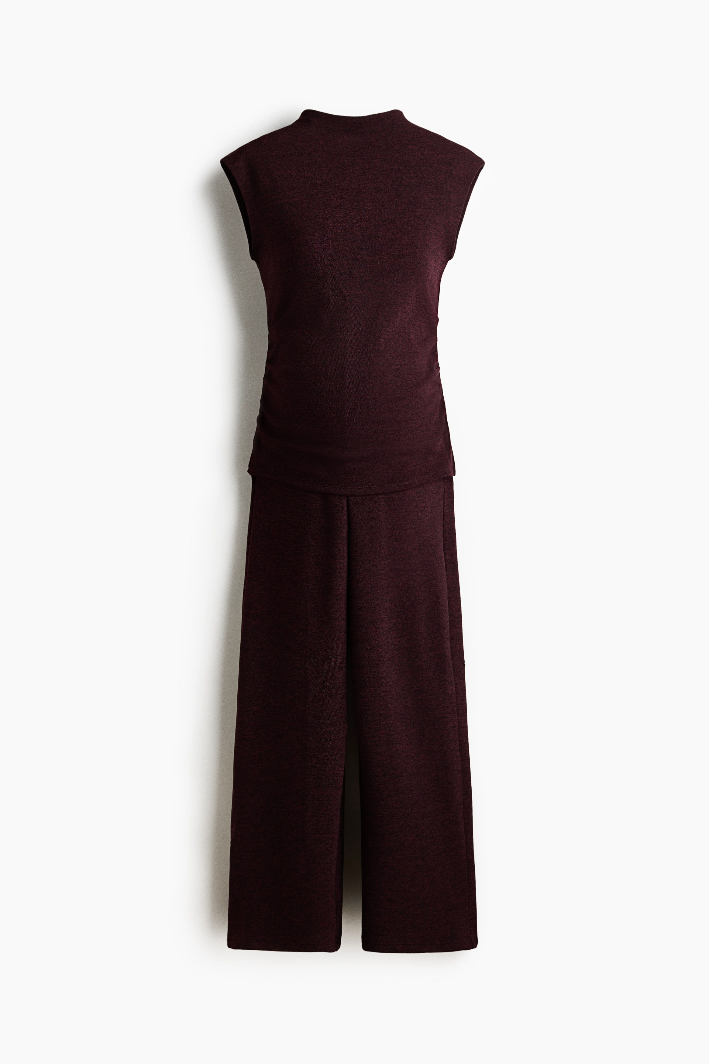 A dark burgundy jersey set, featuring a top with a high neck and cap sleeves and matching trousers, is being worn by the model. The model is sitting on a stool while wearing the set.