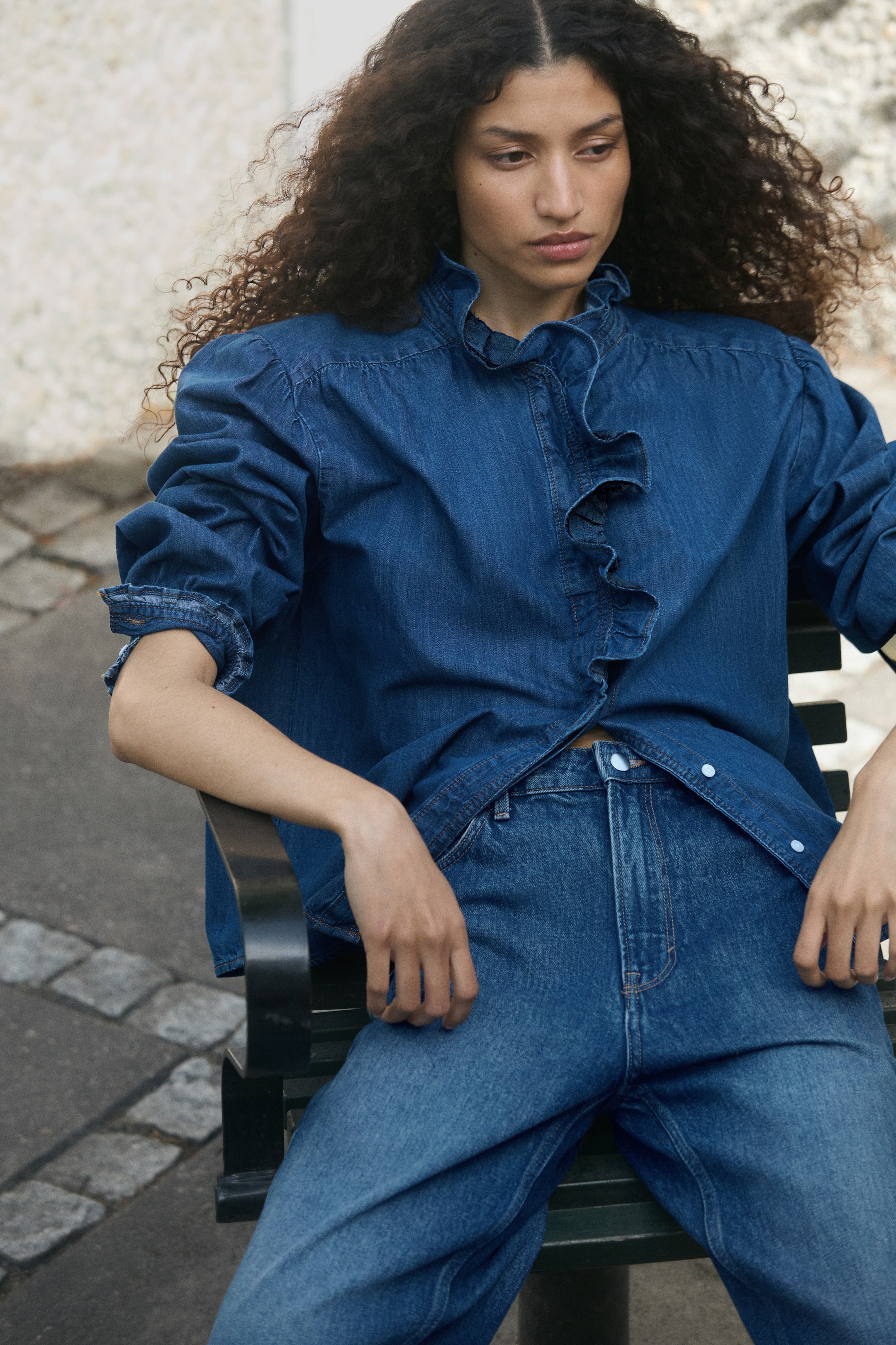 Grotere afbeelding bekijken: Een losvallende katoenen denim blouse in een mediumblauwe wassing wordt gedragen door het model, dat zit. De blouse heeft een hoge halslijn met een gerimpelde ruche-afwerking die doorloopt over de voorzijde van de knoopsluiting, gecombineerd met een bijpassende blauwe denim jeans.