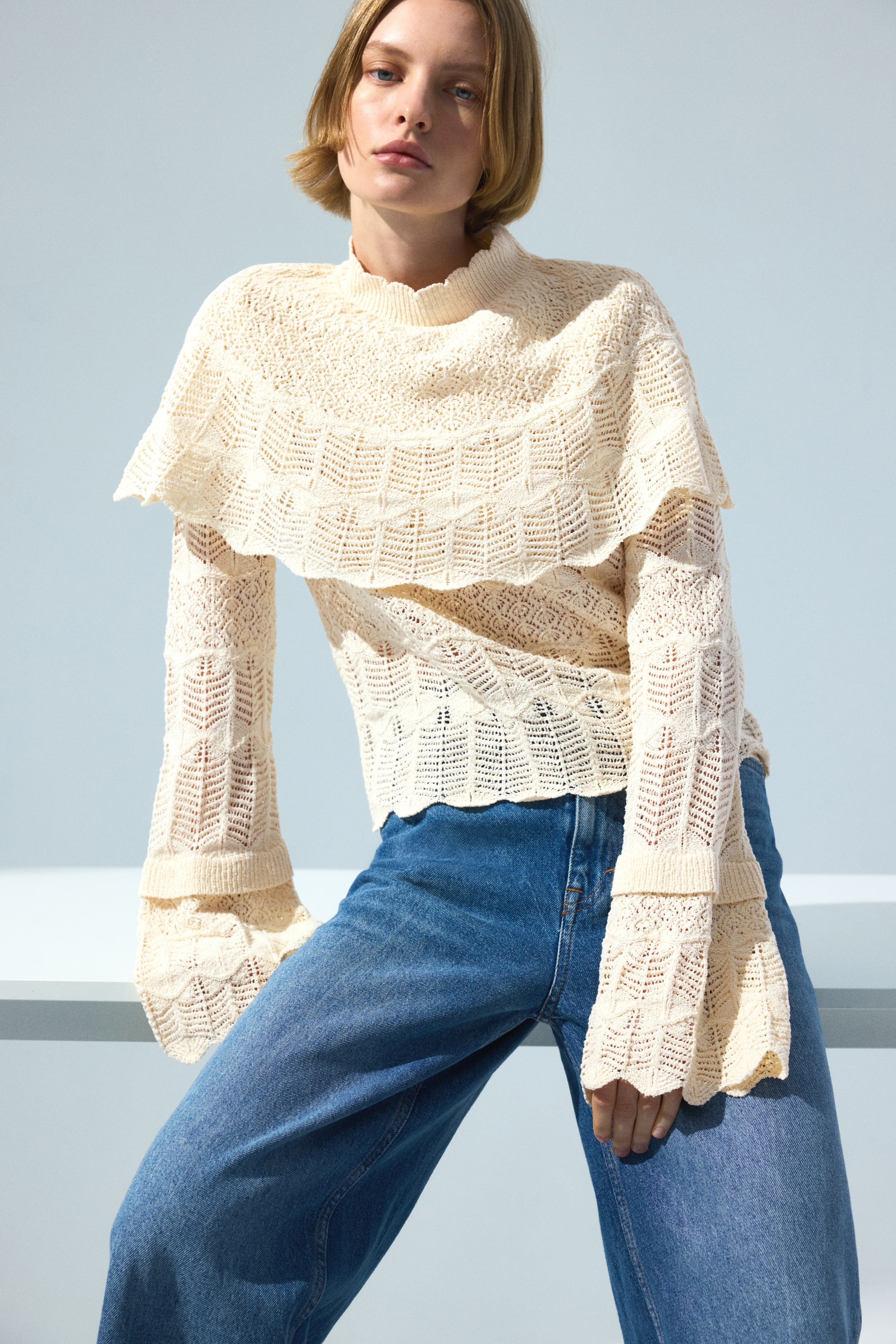 Größeres Bild anzeigen: Ein kurzes Oberteil im Spitzen-Look-Strick wird von dem Model getragen, das auch blaue Jeans trägt. Das Oberteil verfügt über einen runden, gerippten Ausschnitt mit gewelltem Rand und eine darunter befestigte Cape-Schicht. Die langen Ärmel haben einen gerippten Abschluss an den Handgelenken für einen Lagenlook und ausgestellte Manschetten mit gewellten Kanten.