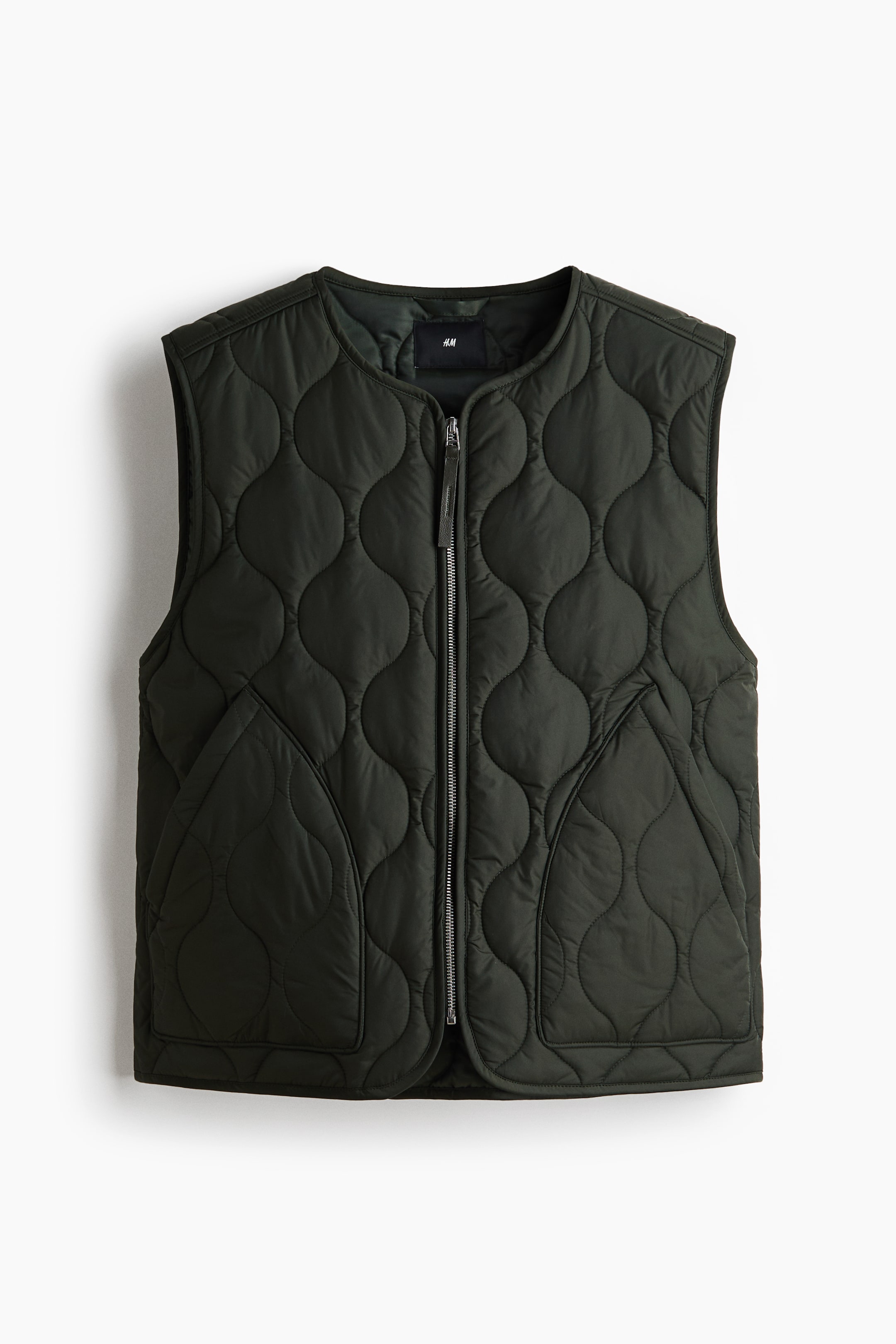 View larger image: A dark green quilted vest is facing forward, featuring a round, collarless neckline and a full-length silver zipper. It is designed with an organic, wavy quilting pattern and two integrated patch pockets on the front.