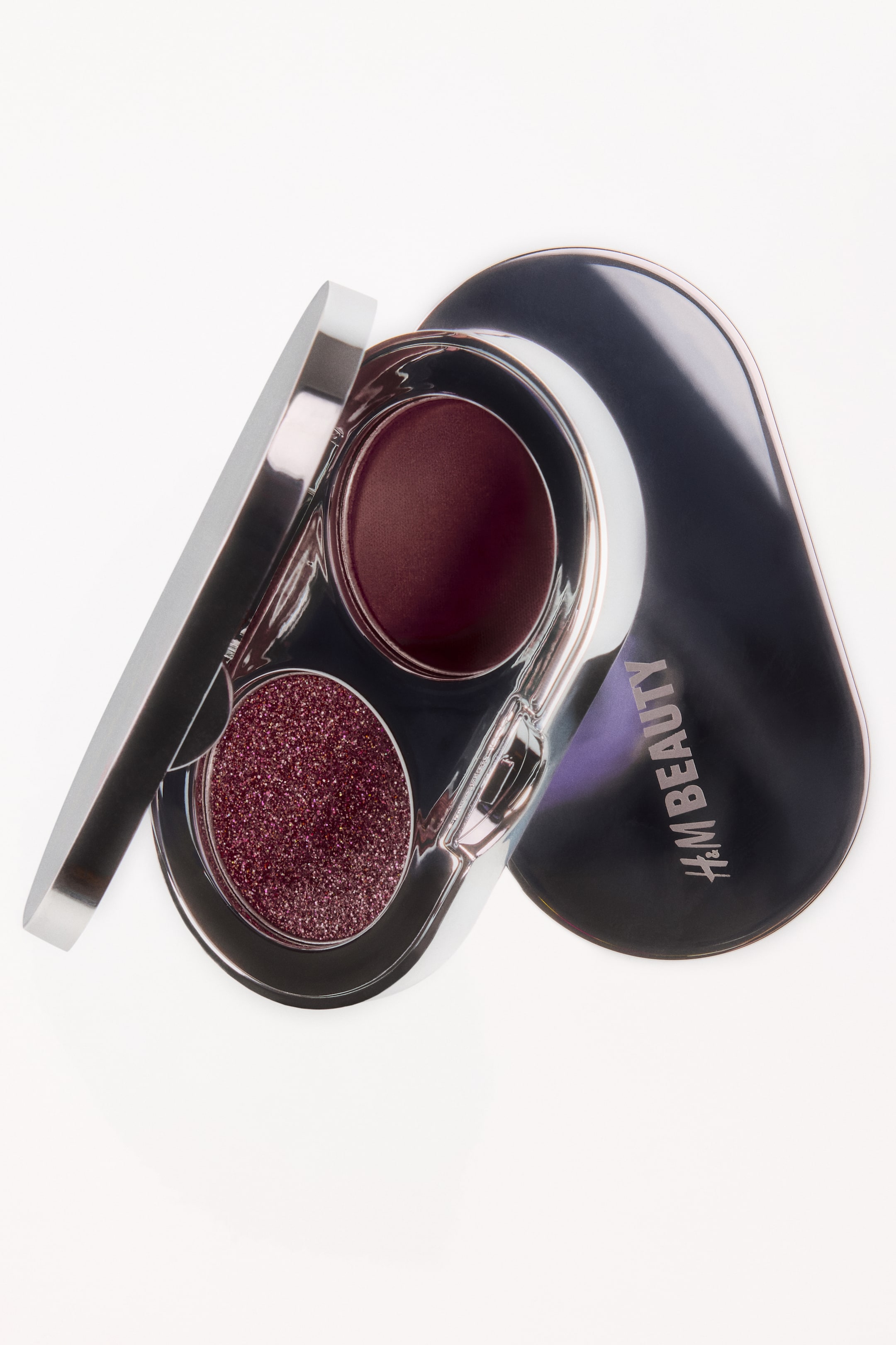 View larger image: An open eyeshadow duo compact is resting at an angle, displaying a metallic silver case with two circular pans containing a deep burgundy satin shade and a sparkling plum glitter topper. The compact is positioned next to its dark metallic lid, which features the "H&M BEAUTY" logo.
