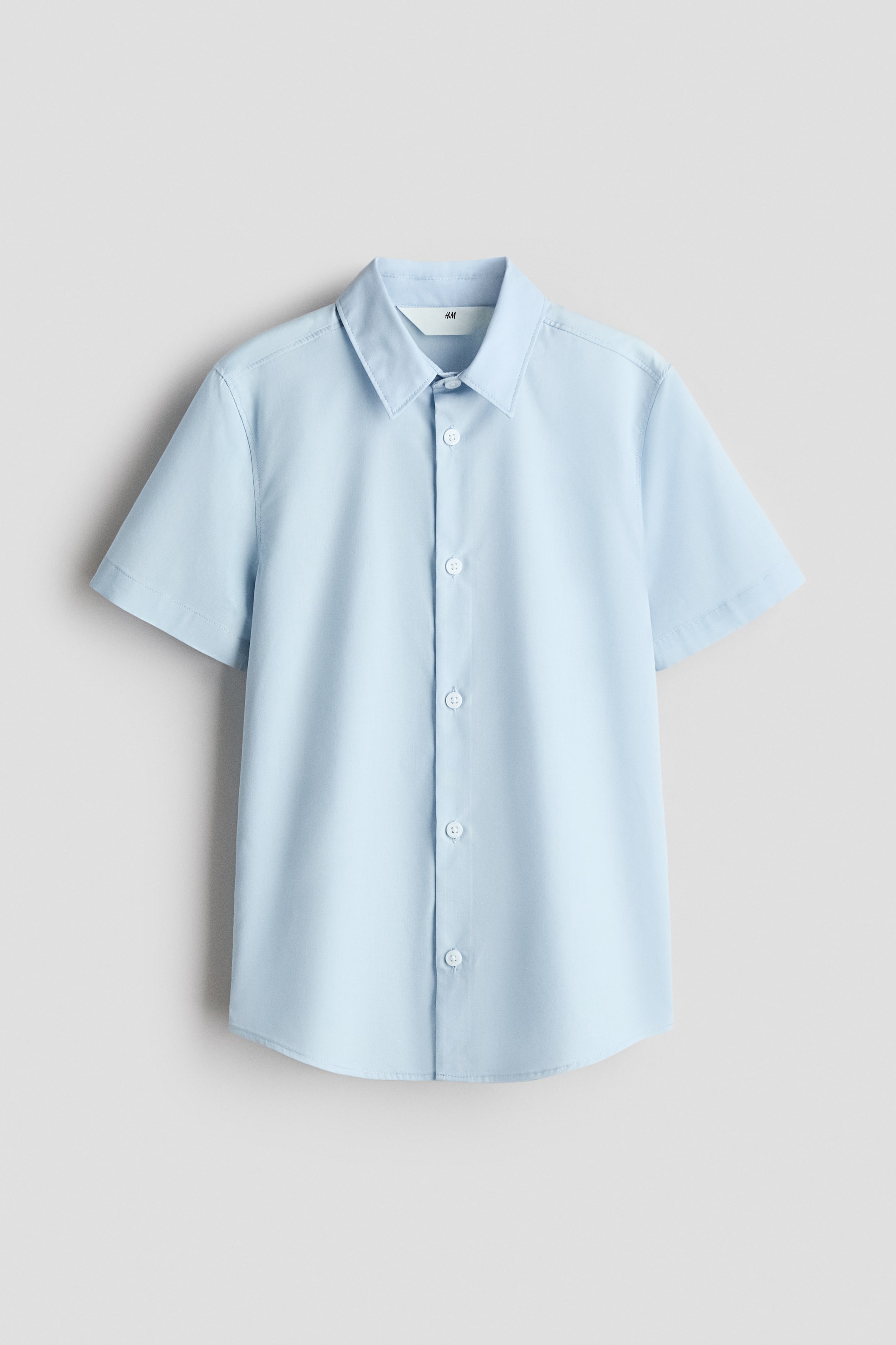 View larger image: A light blue short-sleeve poplin shirt is shown from the front, featuring a collar and a row of white buttons running down the center. It is designed with a rounded hem.