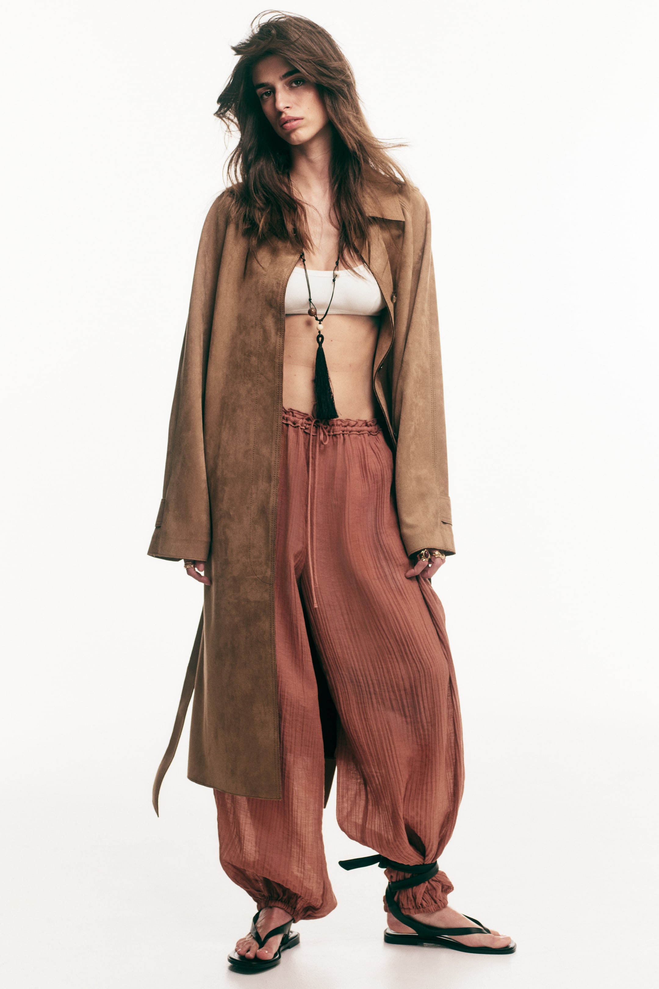 查看大圖: 模特穿著一條紅棕色皺褶織物的寬鬆褲子。這款褲子具有鬆緊抽繩腰部，並飾有窄褶邊，燈籠褲腿配有鬆緊褲腳，搭配黑色涼鞋，涼鞋綁在褲腳外。