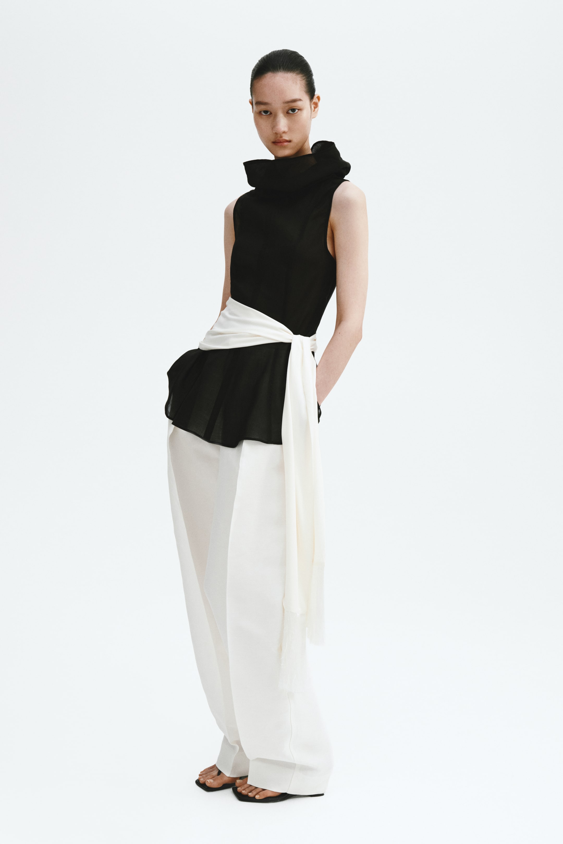 Ver imagem maior: A modelo está a usar um top preto sem mangas. Apresenta uma gola ampla e alta com efeito de cowl e um peplum evasé, e uma faixa branca está atada à cintura.