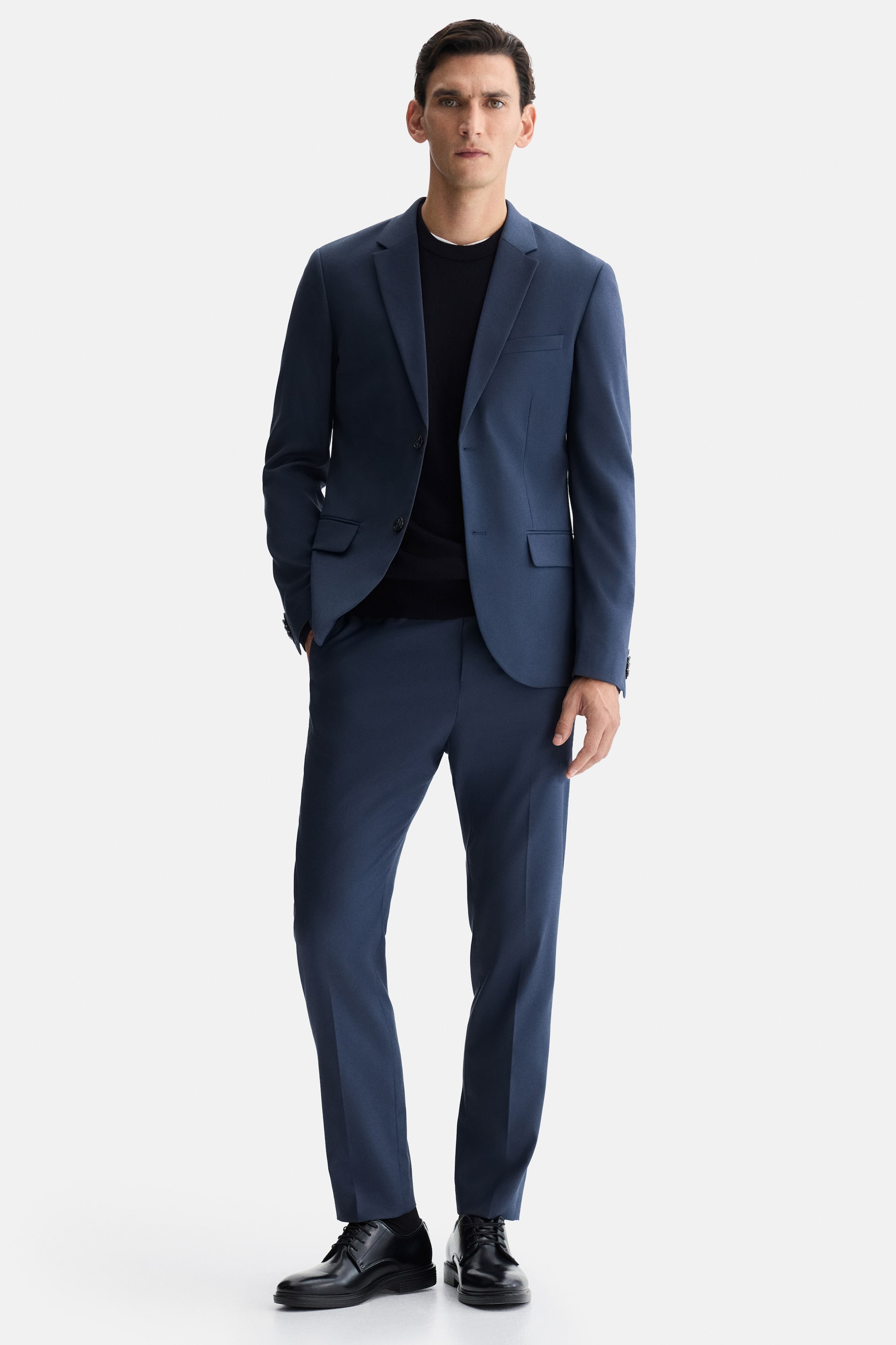 Ampliar la imagen: Un traje azul de dos piezas, que cuenta con un blazer de dos botones con solapas de muesca y bolsillos con solapa, y pantalones rectos a juego, está siendo usado por el modelo. Un suéter negro de cuello redondo está siendo usado debajo del blazer, y zapatos derby negros están siendo usados en los pies.