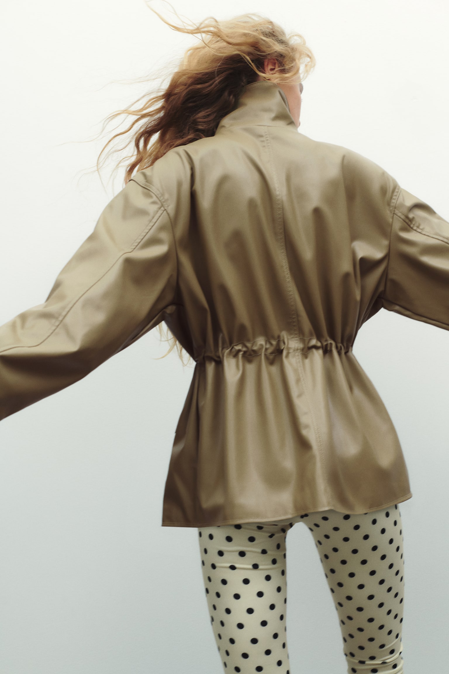 Model mÃ¡ na sobÄ oversized bundu z khaki zelenÃ© povrstvenÃ© lÃ¡tky, zachycenou zezadu. Bunda mÃ¡ vysokÃ½ stojÃ¡Äek, spuÅ¡tÄnÃ¡ ramena a staÅ¾enÃ½ pas na stahovacÃ­ Å¡ÅÅ¯rku, kterÃ½ vytvÃ¡ÅÃ­ nabranÃ½ efekt nad krÃ©movÃ½mi legÃ­nami s puntÃ­ky. - 1