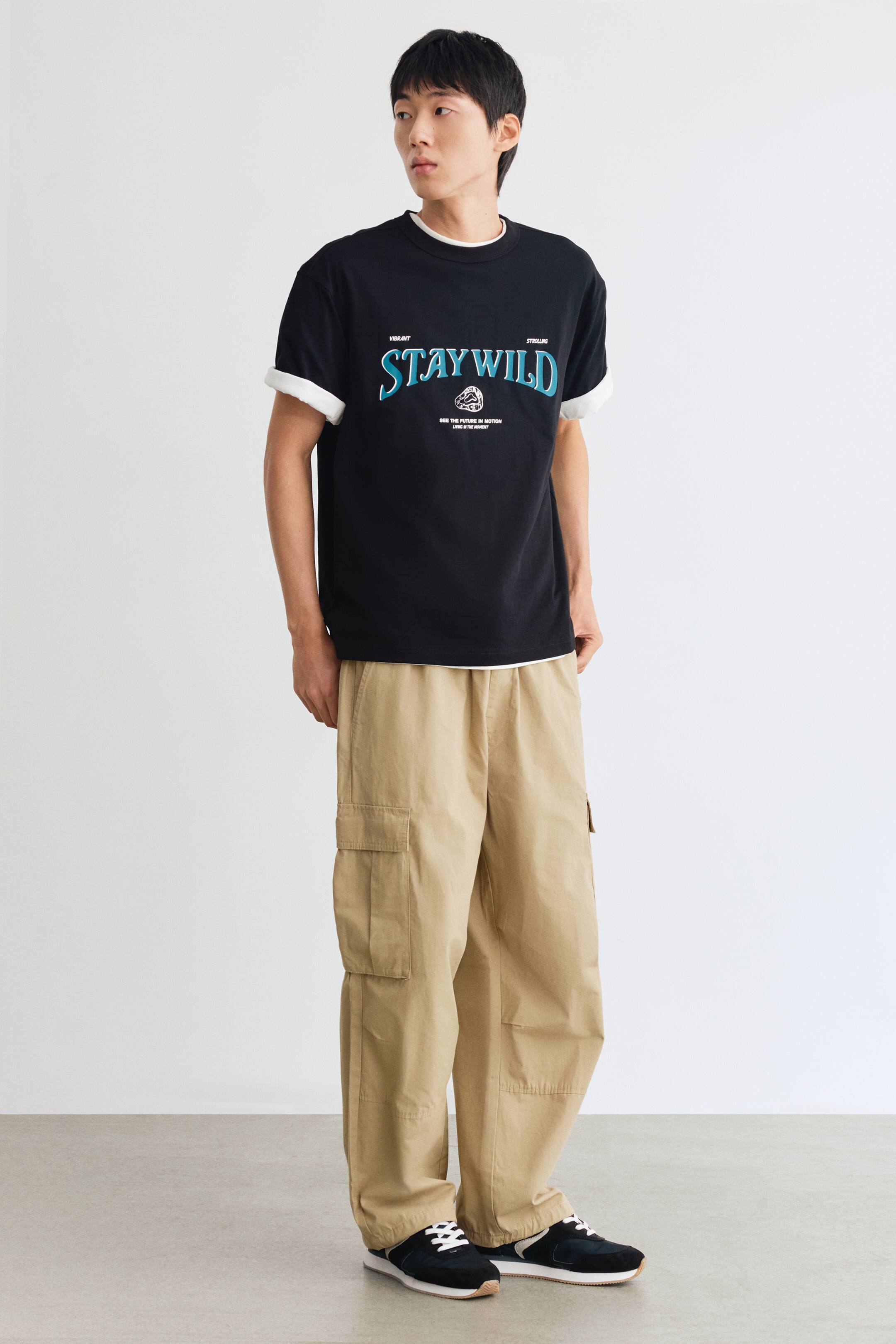 View larger image: A black heavyweight cotton jersey tee featuring a teal and white printed design on the chest is worn by the model. The loose-fit tee is styled over a white T-shirt, visible at the round neckline, rolled sleeves, and hem, and is paired with beige cargo pants.