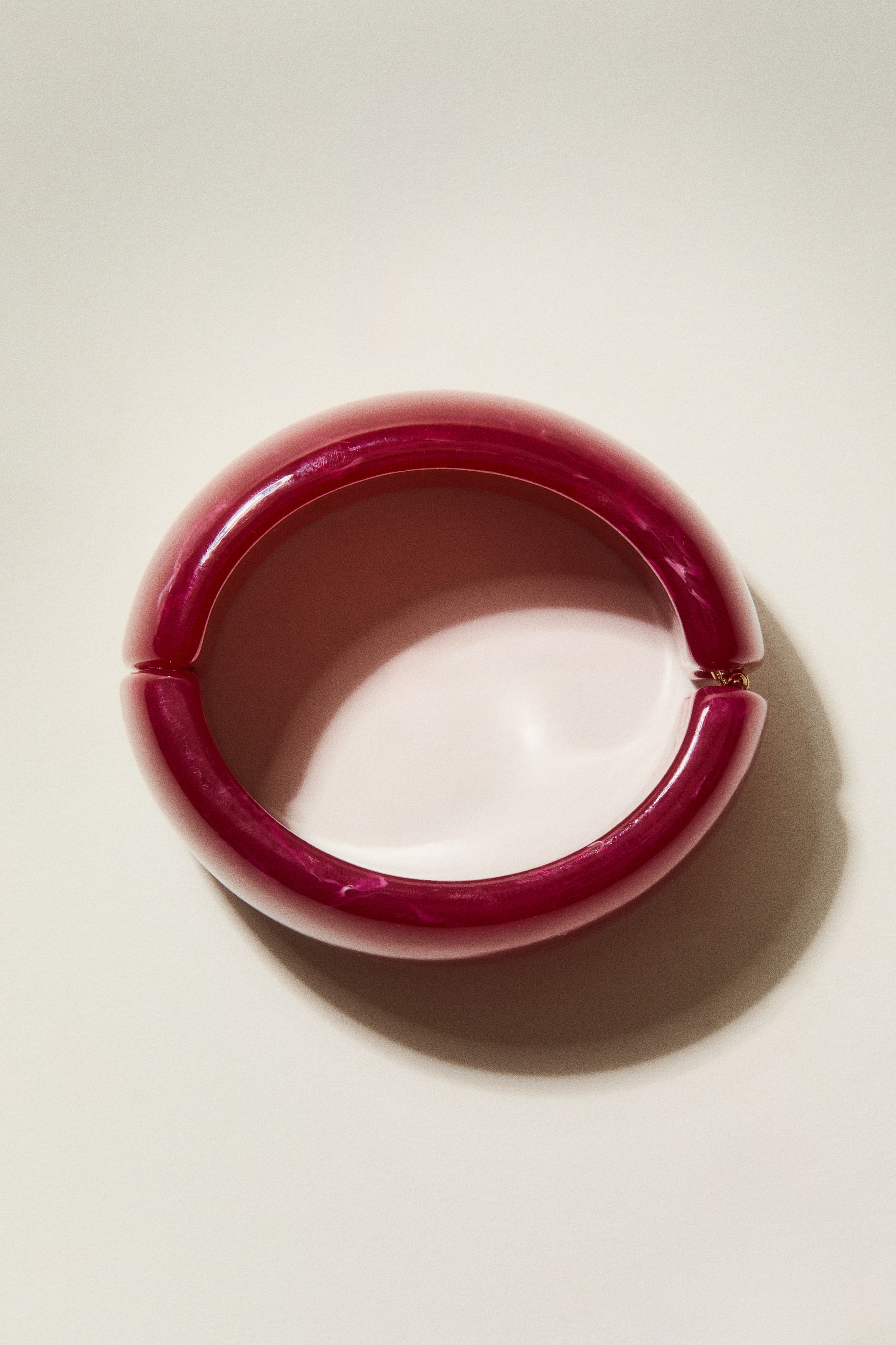 Vis større bilde: Et kraftig, hengslet armbånd viser en oval form fra forsiden, laget av blank, marmorert mørk rosa plast med lysere virvler. En liten, gullfarget hengemekanisme forbinder de to halvdelene på høyre side.