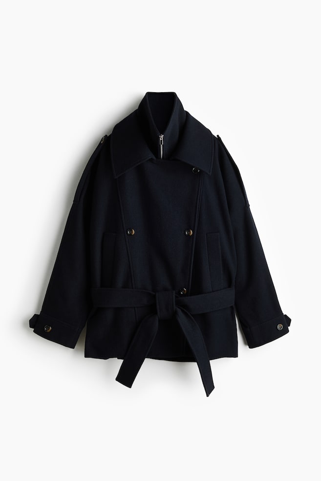 A short, double-breasted navy coat is shown from the front, featuring a double-layered collar design with a zippered inner funnel neck and a large outer collar. The oversized coat features dropped shoulders with buttoned epaulettes, adjustable button tabs at the cuffs, and a matching fabric belt tied at the waist. - 1