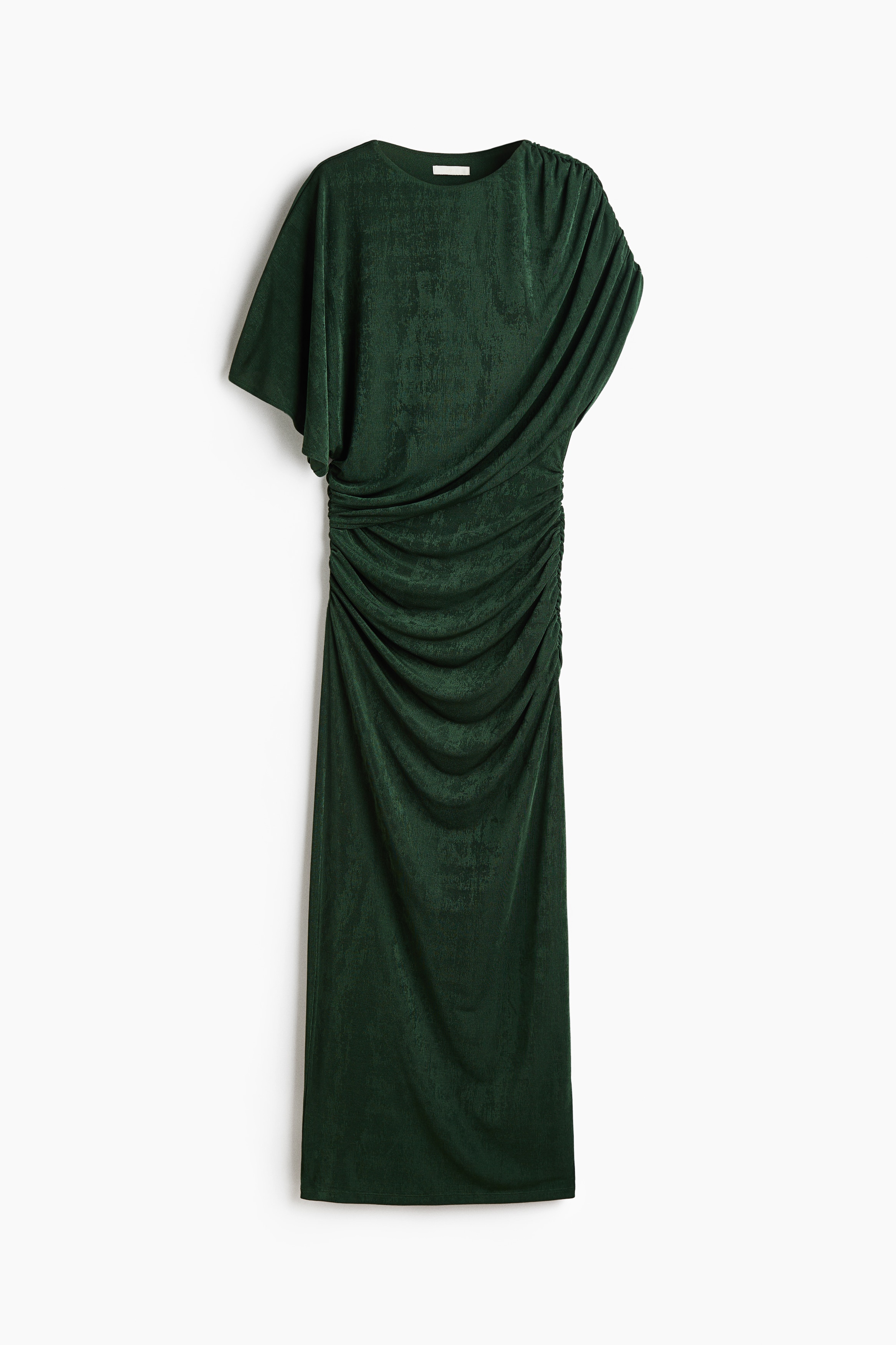 The model is wearing a dark green fitted stretch jersey dress with a round neckline and gathered seams that create a draped effect across the body. The asymmetrical design features a draped, cape-like sleeve on one side and a short sleeve on the other.