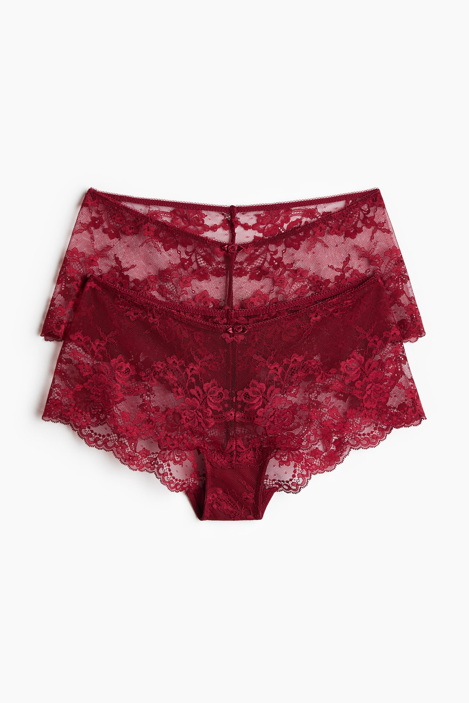 Two pairs of dark red lace underwear are lying flat, stacked one slightly behind the other and facing front. The underwear feature a sheer floral lace design with scalloped trim along the edges and a small decorative bow at the centre of the waistband.