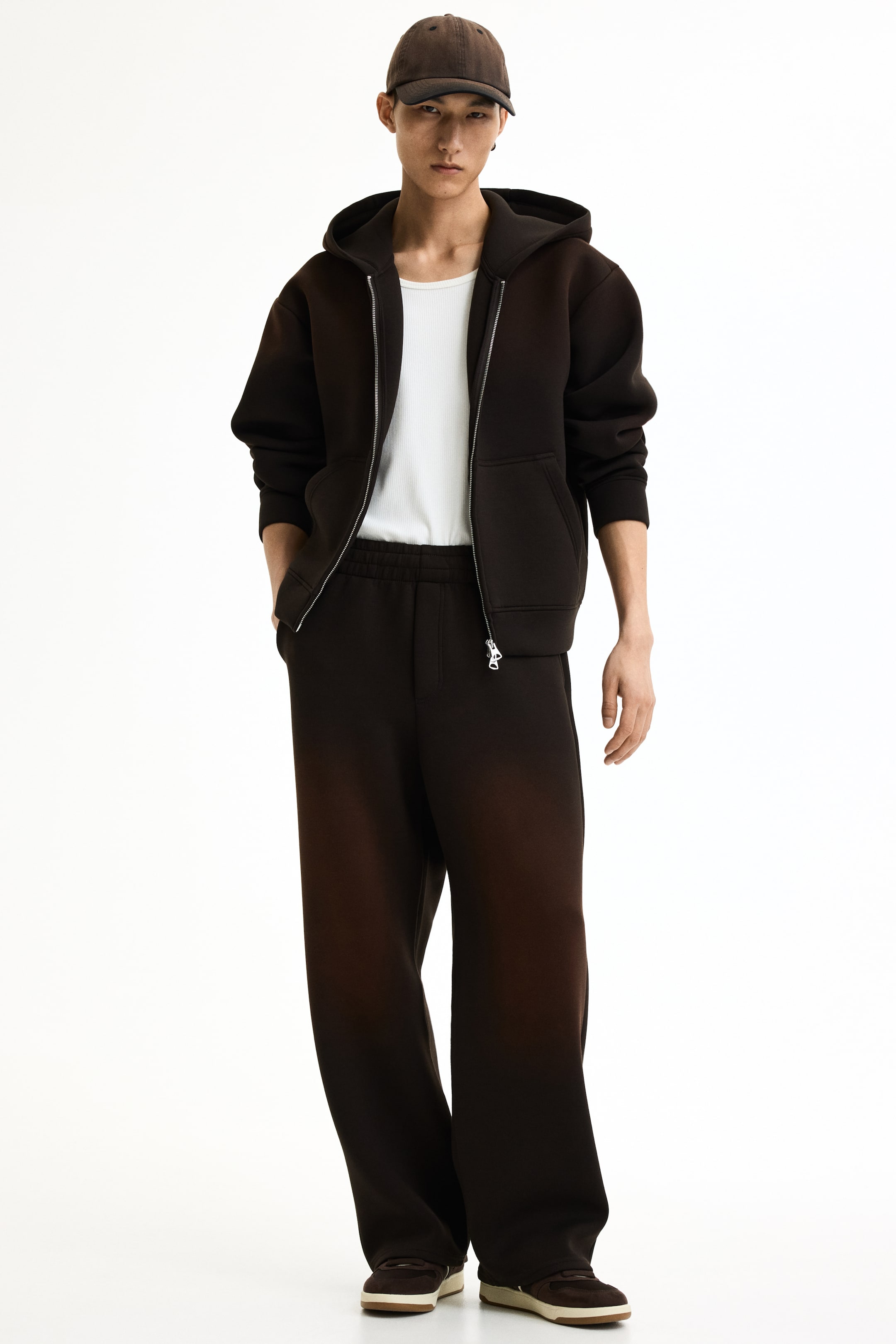 View larger image: A dark brown full-zip hoodie and matching wide-leg sweatpants are showcased on a model facing slightly to the right. The hoodie features a silver-toned zipper and front kangaroo pockets, worn open over a white ribbed undershirt. The sweatpants highlight an elastic waistband and a relaxed fit, pooling at the ankles.
