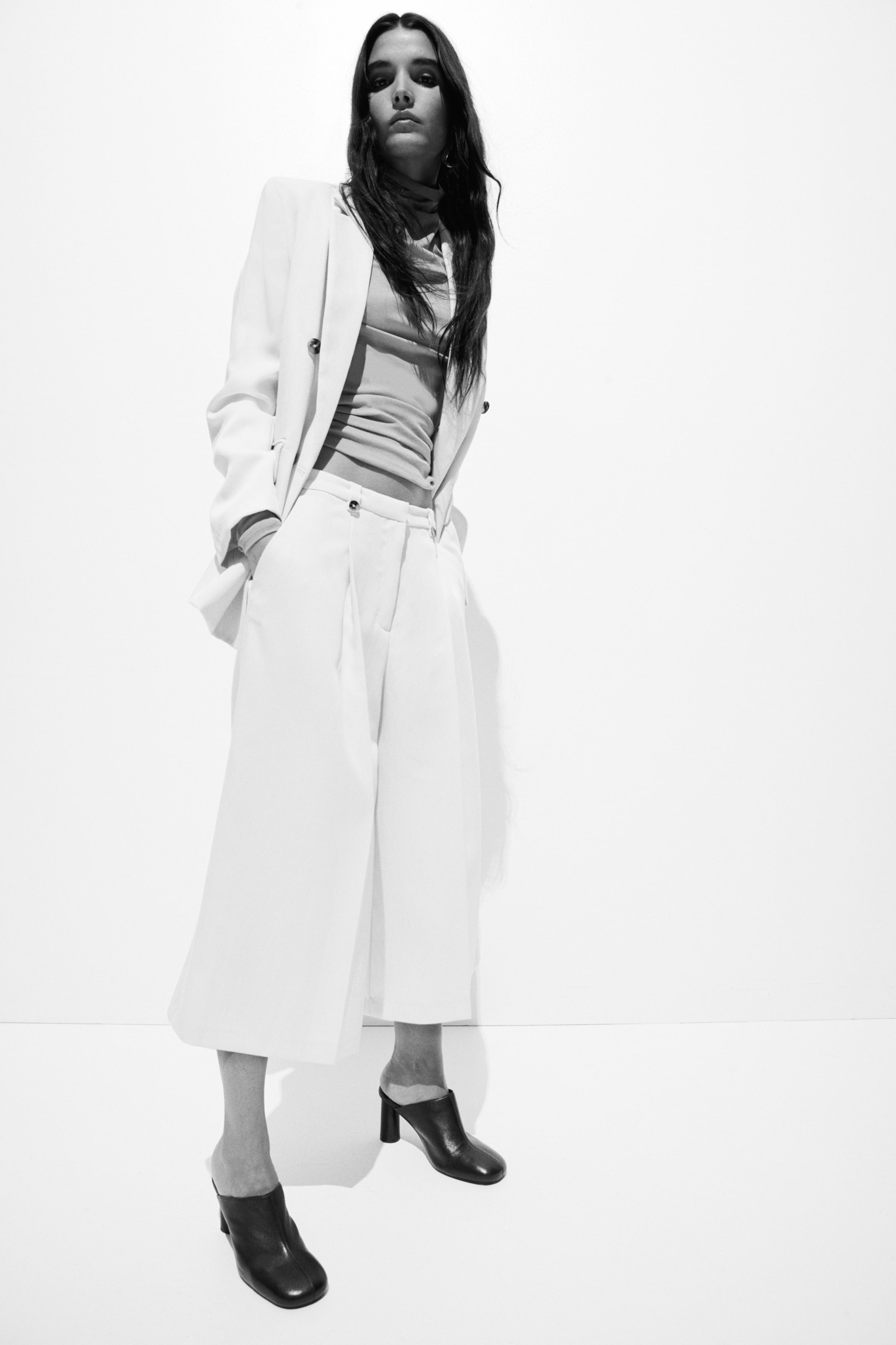 View larger image: A white double-breasted blazer with notch lapels and dark buttons is styled open over a light-coloured, ribbed, high-neck, long-sleeved top. It is teamed with white, wide-leg, pleated, cropped trousers featuring a dark button fastening, and dark mule-style shoes with block heels and square toes complete the look. The entire ensemble is showcased from a low angle.