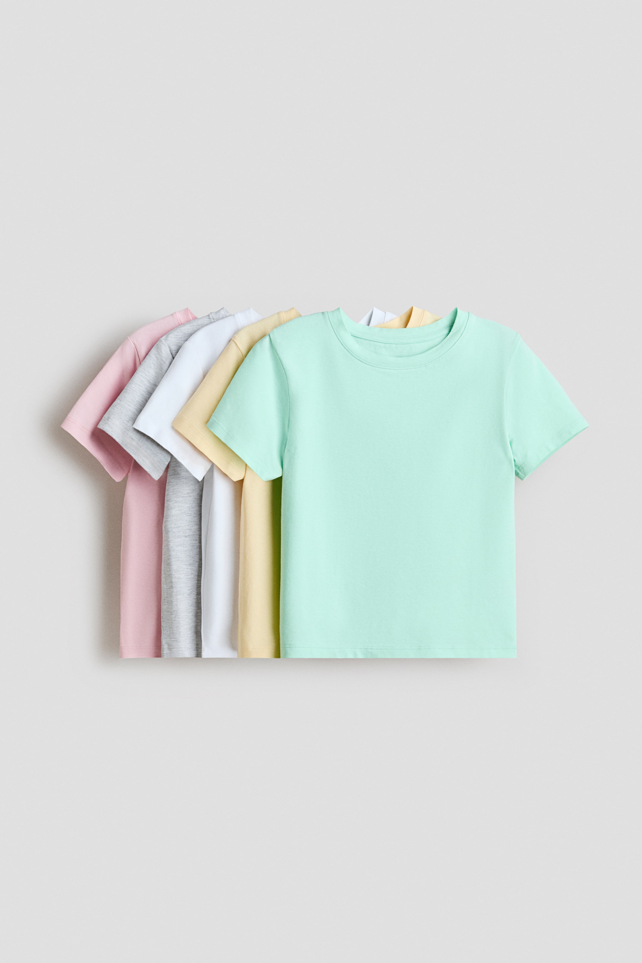 Grösseres Bild ansehen: T-Shirts aus weichem Baumwoll-Jersey werden von vorne in einer fächerförmigen Anordnung präsentiert. Jedes T-Shirt verfügt über einen gerippten Besatz am Halsausschnitt, sanft fallende Schultern und kurze Ärmel und ist in Farben wie Mintgrün, Gelb, Weiß, Grau und Rosa erhältlich.