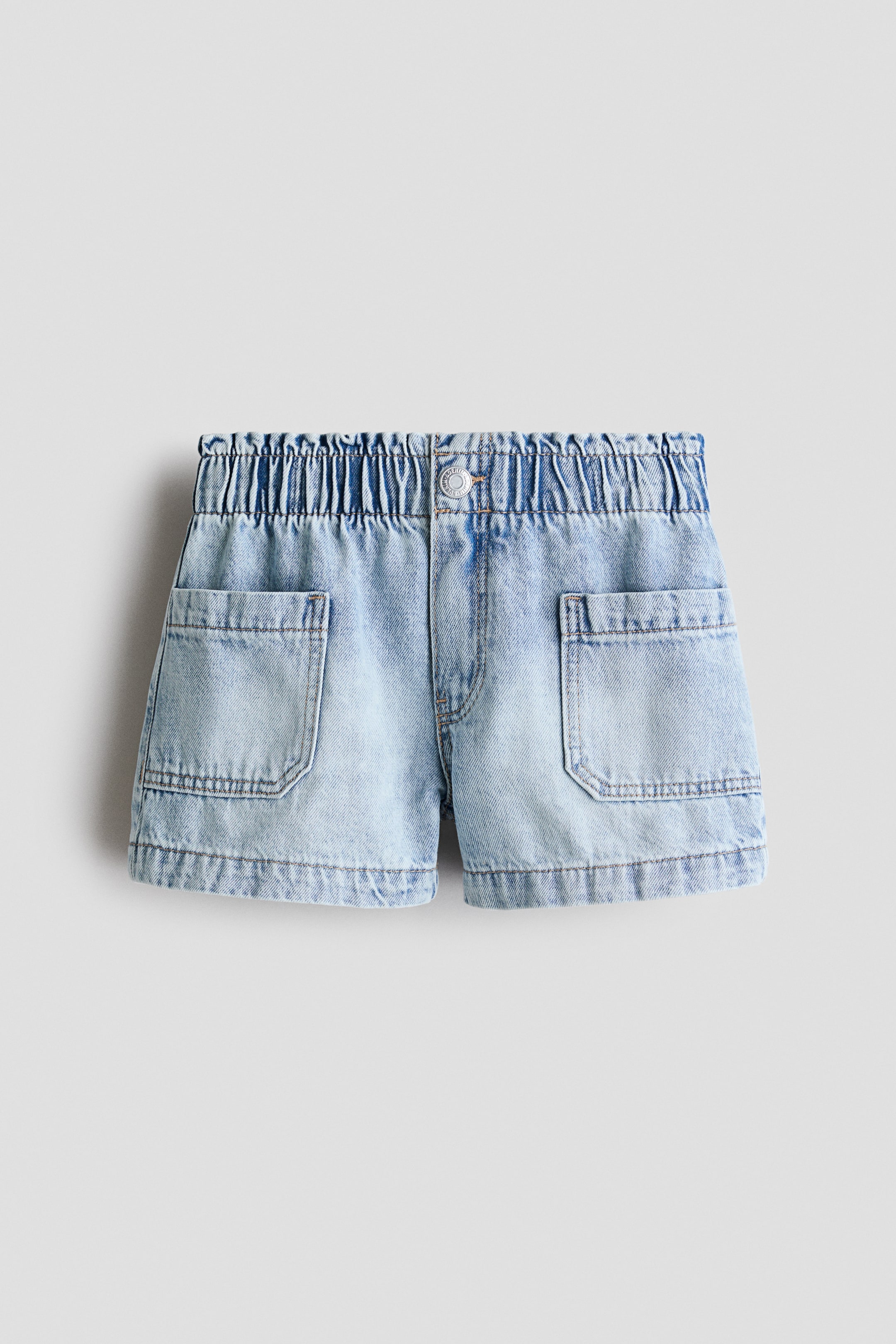 View larger image: Loose-fit shorts in washed light blue cotton denim are shown from the front, featuring an elasticated, gathered paper bag waist, a faux fly with a single snap closure, and two large rectangular patch pockets on the front.