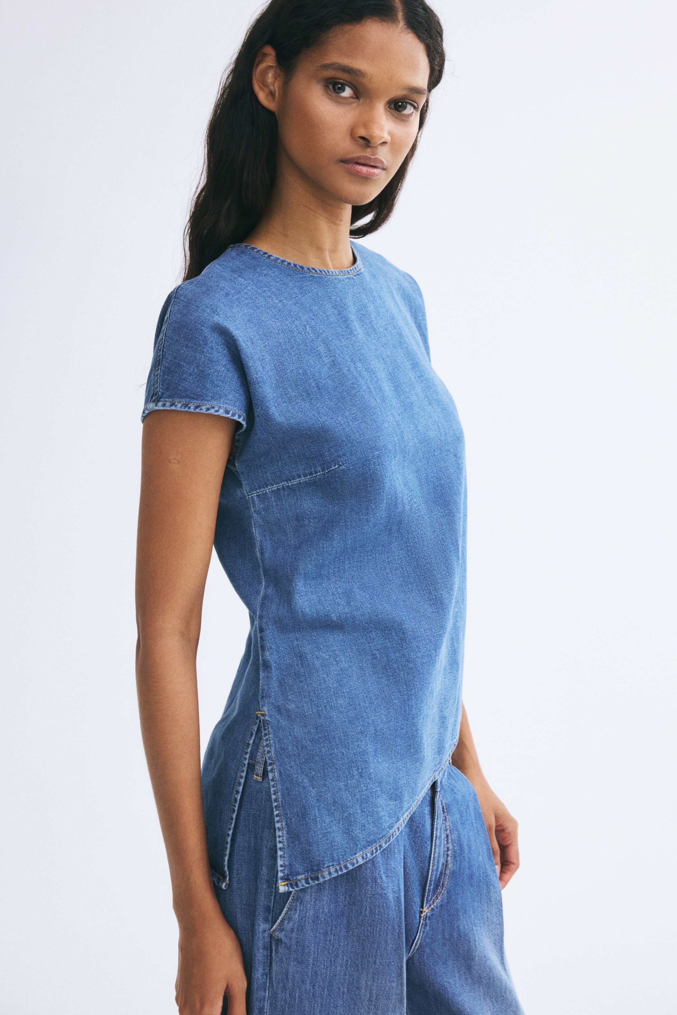 A blue denim top is shown from the front, featuring a crew neck, extended cap sleeves, and a pronounced asymmetrical hemline sloping upward from left to right. The medium-wash top displays subtle bust darts for shaping and contrast topstitching along the seams.
