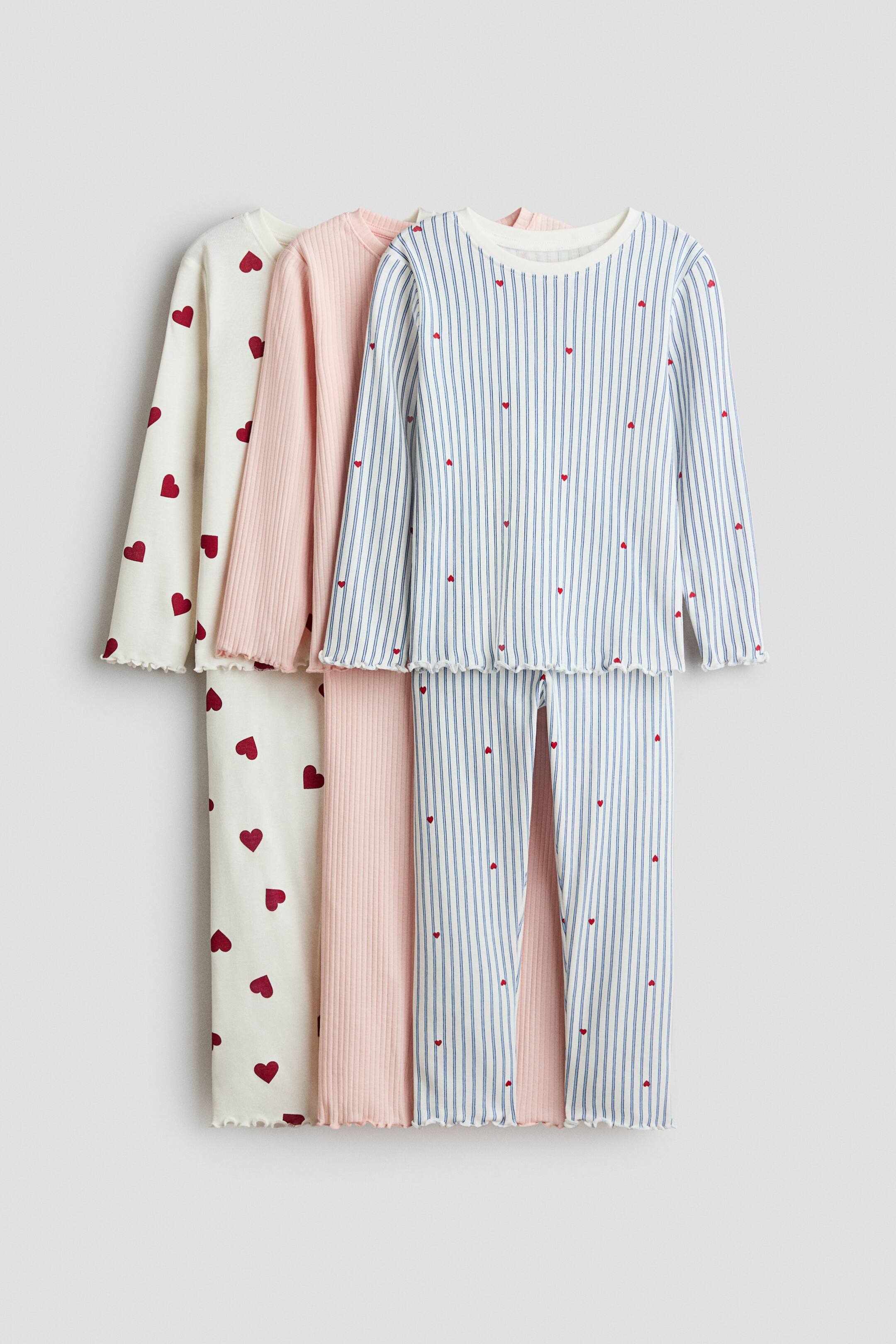 View larger image: Three sets of children's pyjamas are hanging vertically, slightly overlapping. The leftmost set is an off-white top and bottom featuring a red heart print, the middle set is a solid light pink ribbed top and bottom, and the rightmost set is a white top and bottom with blue vertical stripes and small red heart accents; all pieces are displaying long sleeves, long trousers, round necklines, and lettuce-edge hems.