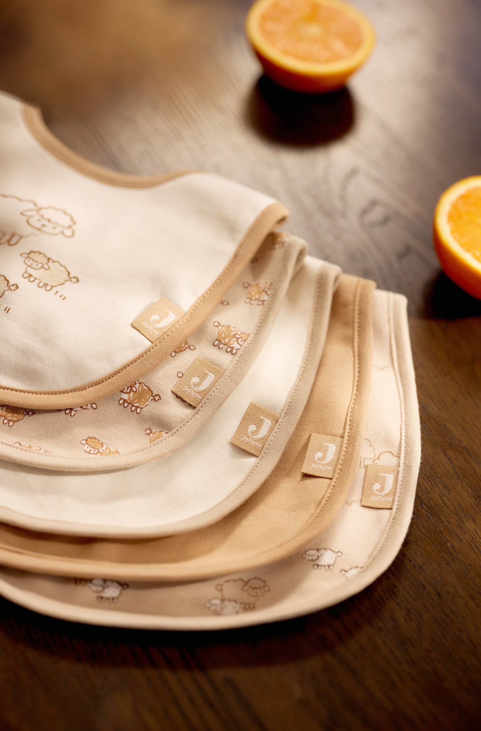Een set van vijf slabbetjes voor babyâs in neutrale tinten crÃ¨me, beige en zandkleur is in een diagonale opstelling overlappend gerangschikt. Het voorste slabbetje toont een cartoonillustratie van een schaapsgezin met een zandkleurige bies, terwijl de onderliggende slabbetjes effen neutrale kleuren en all-over schaapjesprints hebben, met halsluitingen met klittenband.