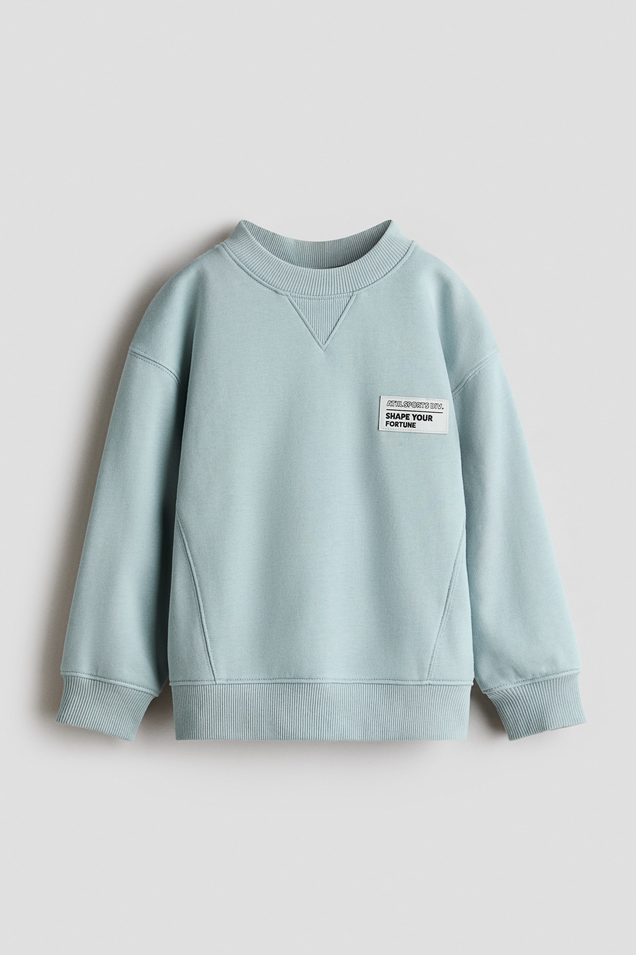 Visa större bild: Den ljusblåa sweatshirten har en ribbad rund halsringning med V-söm detalj, långa ärmar med ribbade muddar och en ribbad nederkant. En vit rektangulär lapp med texten 'ATHL. SPORTS DIV. SHAPE YOUR FORTUNE' i svart text är placerad på vänster bröst.