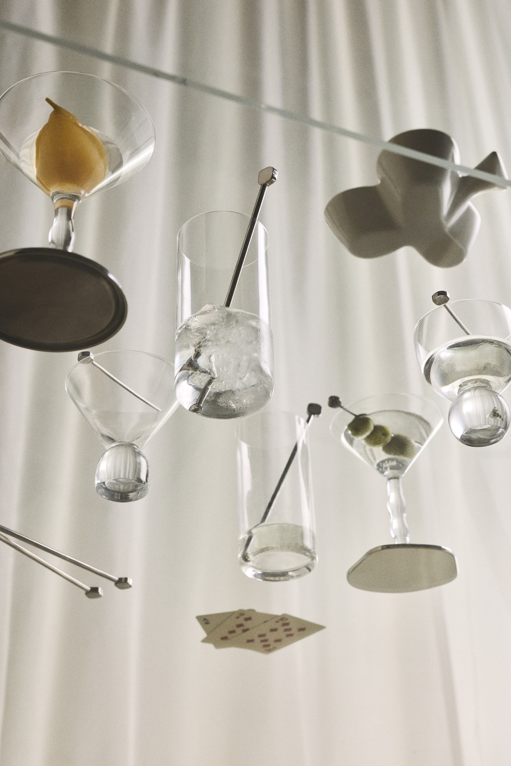 View larger image: Stainless steel cocktail picks and drink stirrers featuring small, geometric tops with softly rounded edges are resting inside various clear cocktail and highball glasses, viewed from below. One of the sleek picks is skewered with olives inside a martini glass.