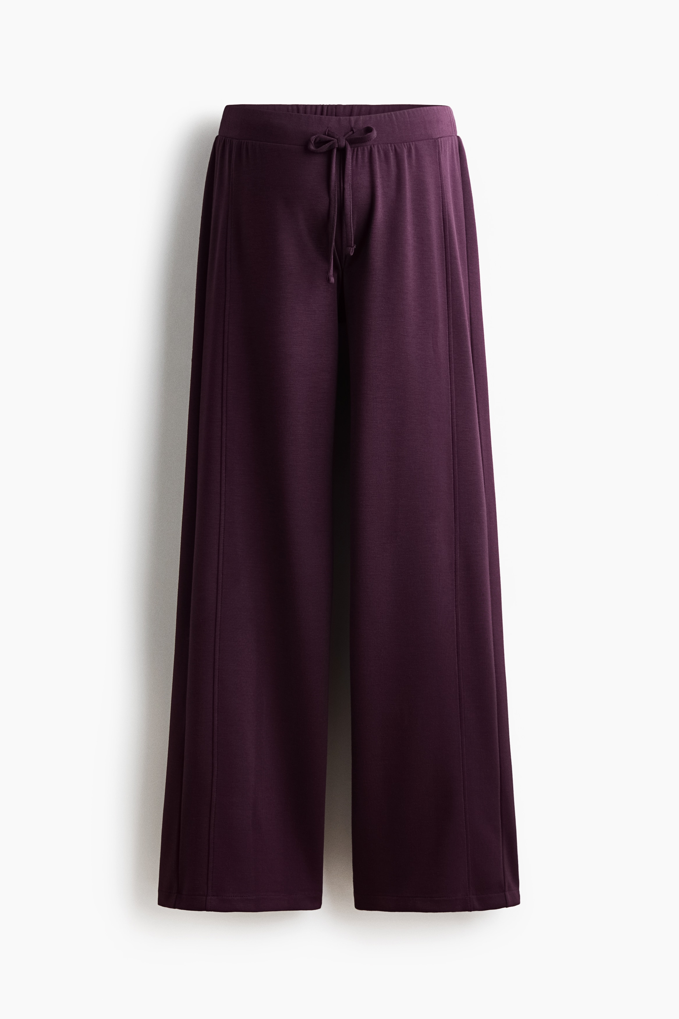 Dark purple wide-leg trousers featuring a visible front seam and a narrow drawstring waist are worn by the model. The relaxed-fit trousers are styled with a white tank top and an open white button-up shirt, highlighting the low, elasticated waistline.